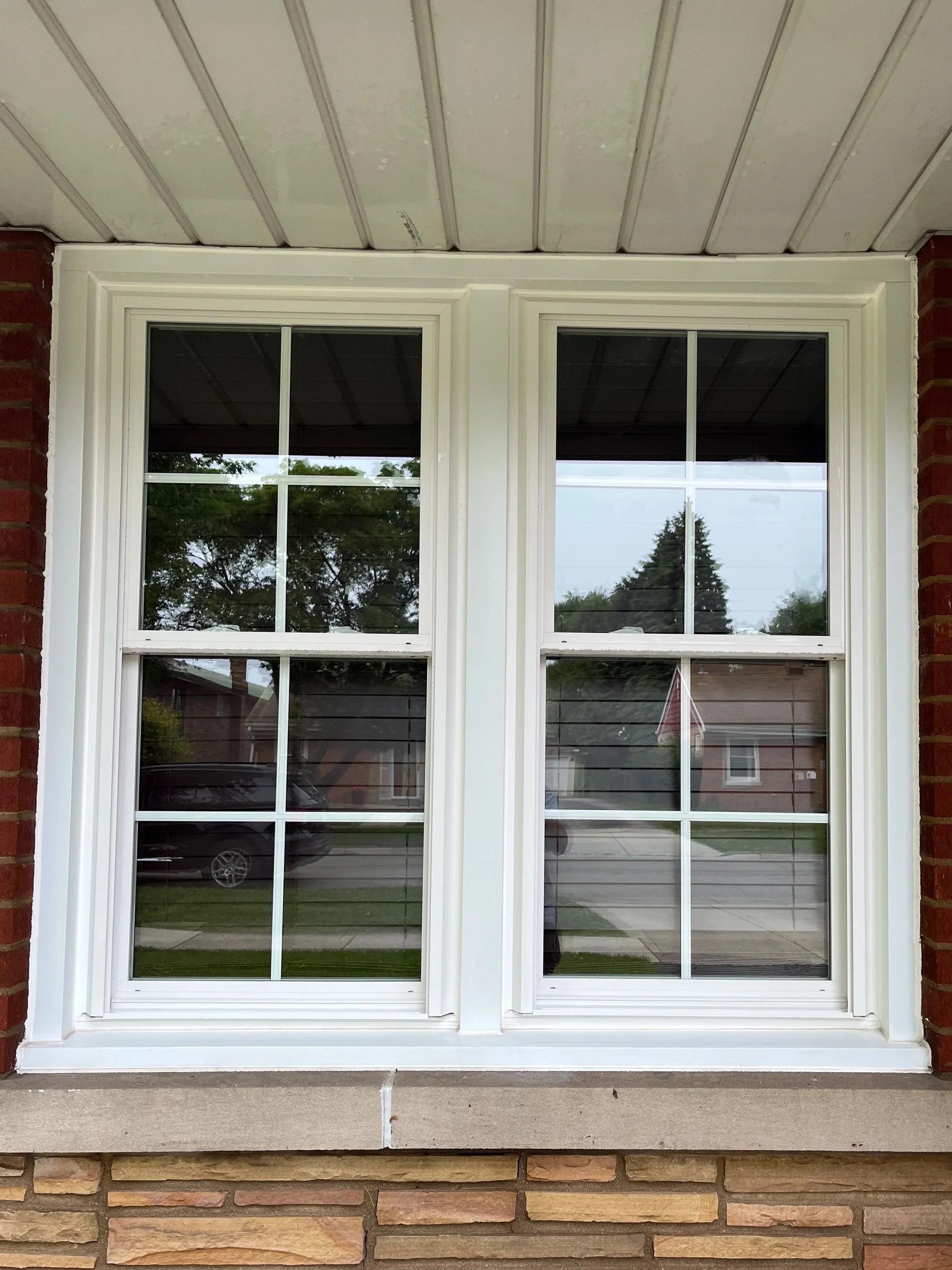 Double-hung window with white framing on a brick house exterior, reflecting trees and neighboring houses.
