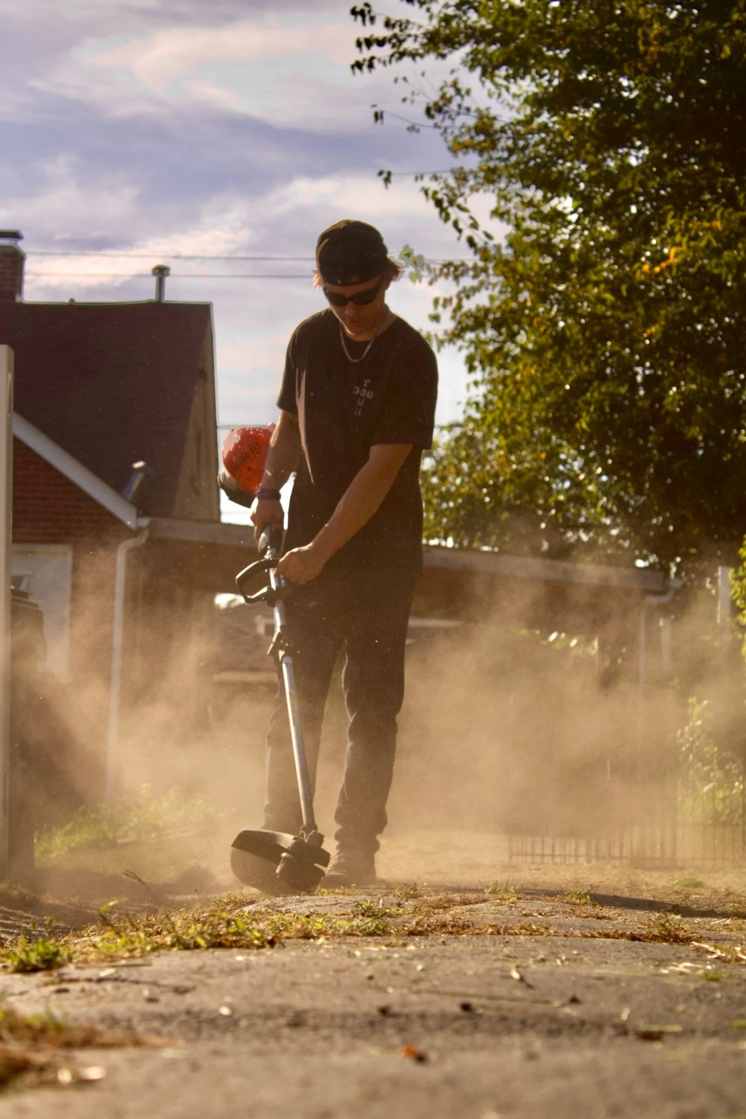 A person wearing sunglasses, a black baseball cap, and a black T-shirt riding a scooter on a dusty sidewalk, with trees and houses in the background during sunset.