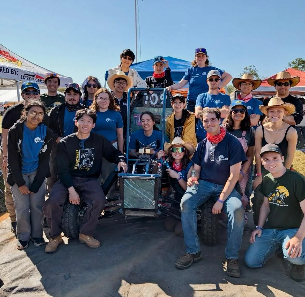 Group of young people gathered outdoors around a robotic vehicle at Baja SAE Arizona competition, smiling and posing for the photo, under tents on a sunny day.