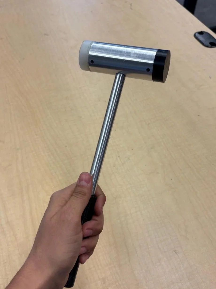 A hand holding a metal mallet with a rubber handle over a light wooden table.
