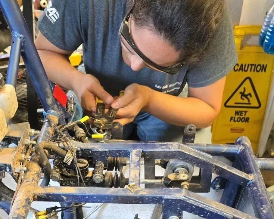 A woman works on repairing a mechanical vehicle in a workshop, wearing safety glasses and a gray t-shirt, with a yellow wet floor caution sign visible in the background.