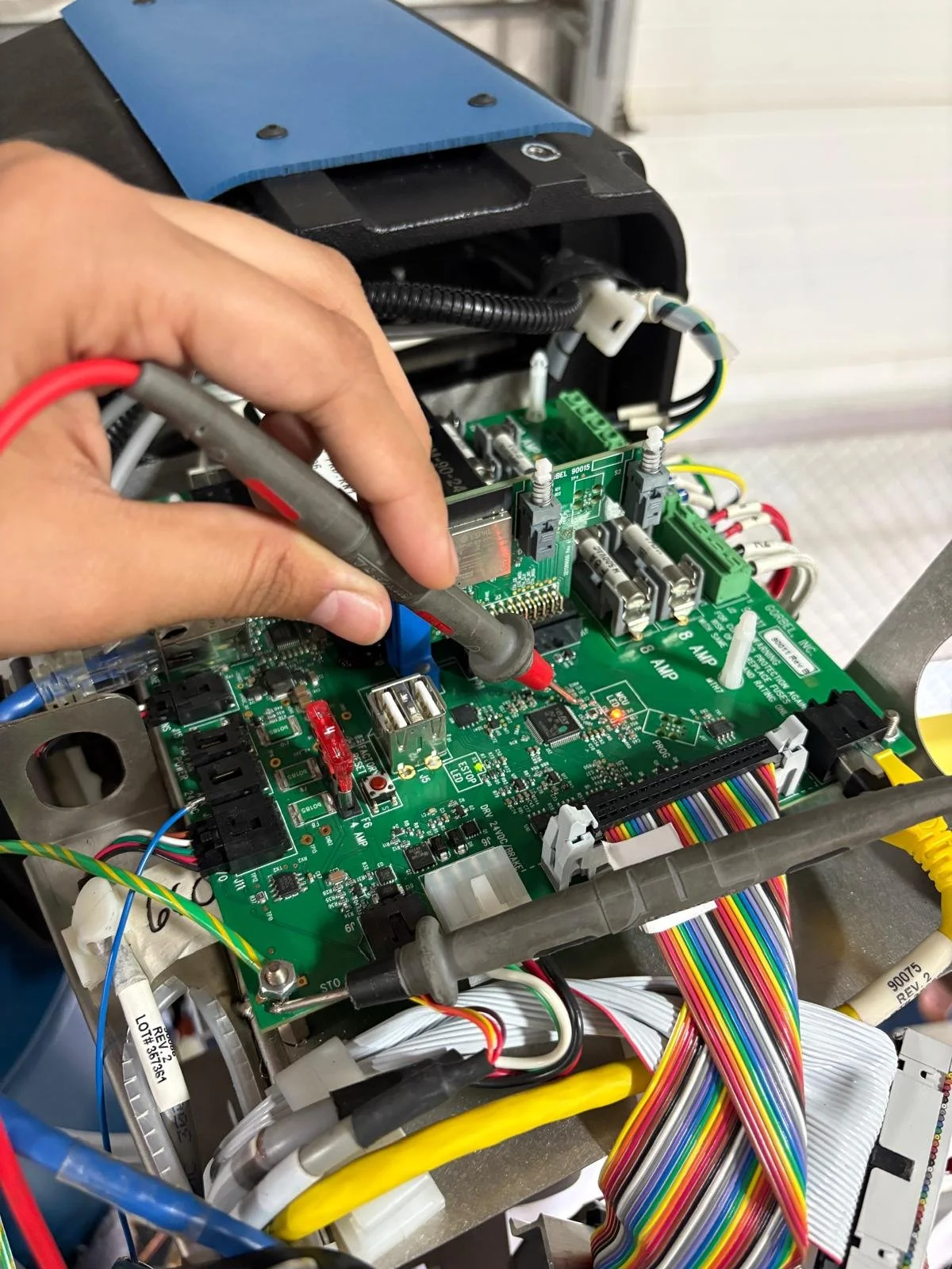 Close-up of a person's hand testing a green printed circuit board (PCB) with a multimeter, surrounded by colorful ribbon cables and electronic components.