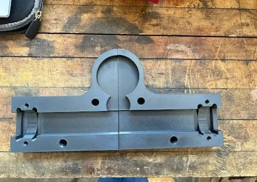 Gray metal mechanical part with multiple holes and a circular cutout, placed on a wooden surface.