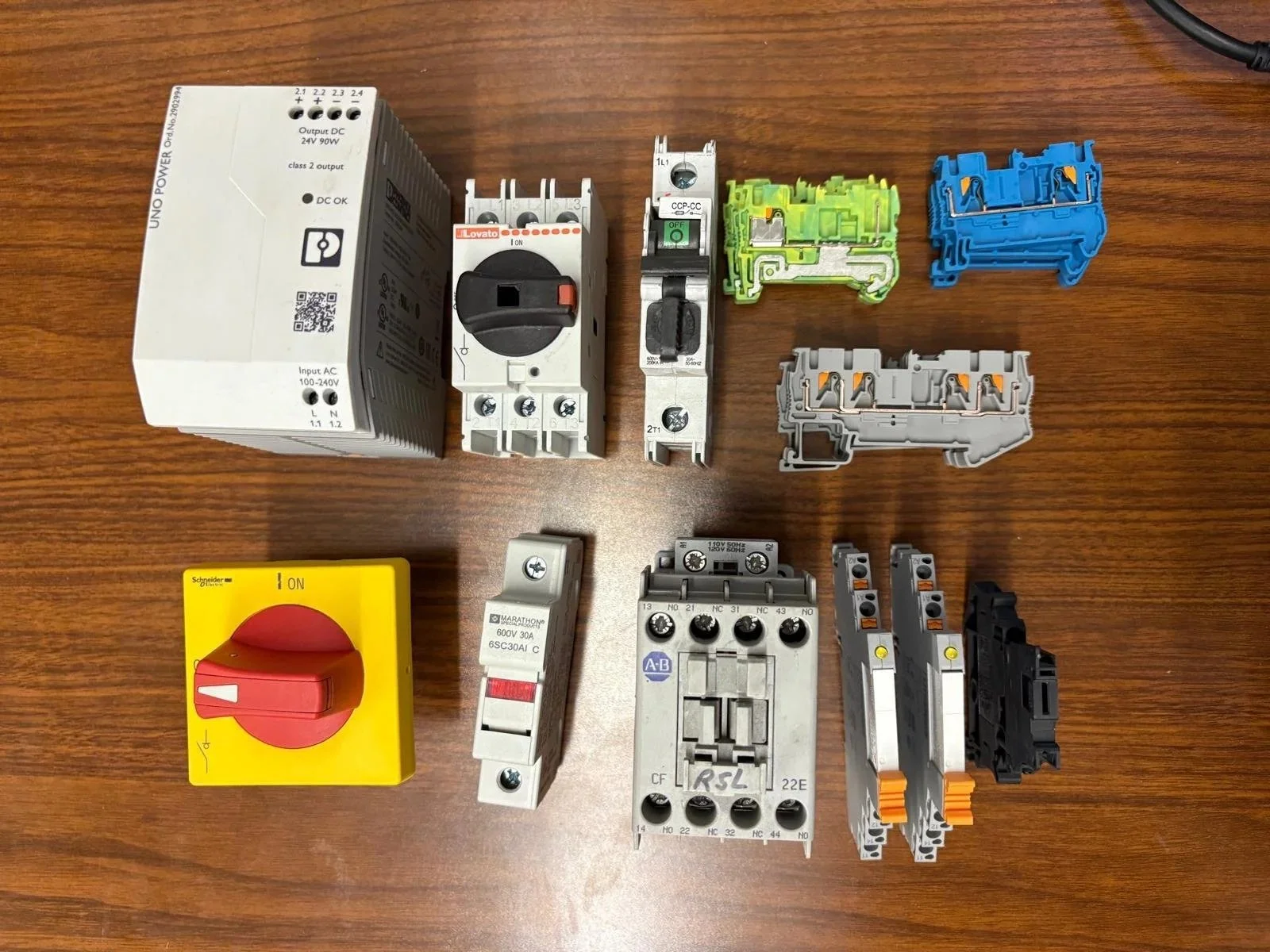 Collection of electrical components on a wooden surface, including a large white power supply, circuit breakers, terminal blocks in blue, green, white, grey, a yellow switch, and other electrical connectors.