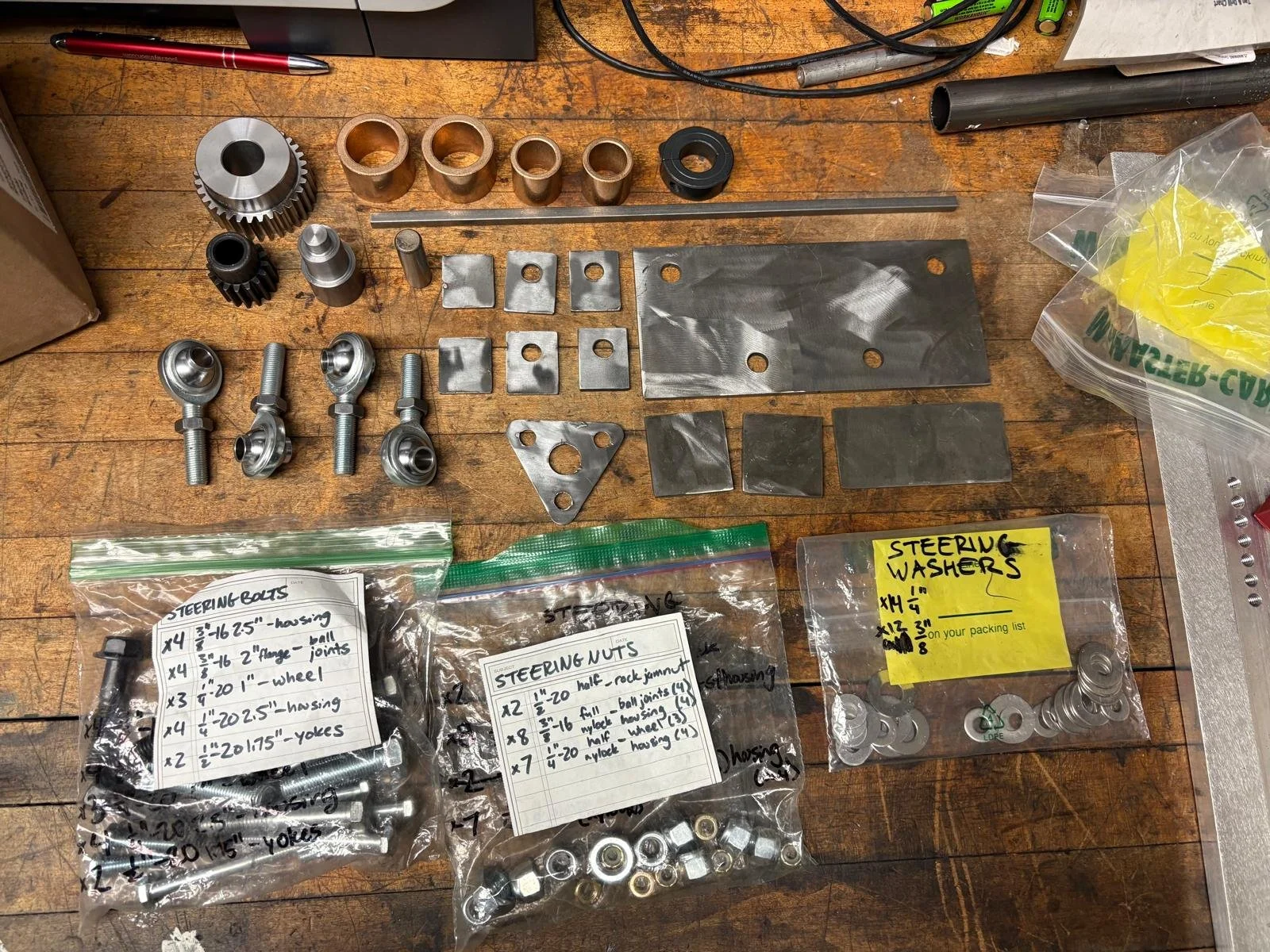 Assorted mechanical parts, including bolts, gears, brackets, and washers, arranged on a wooden workbench with handwritten labels and packaging in an industrial setting.