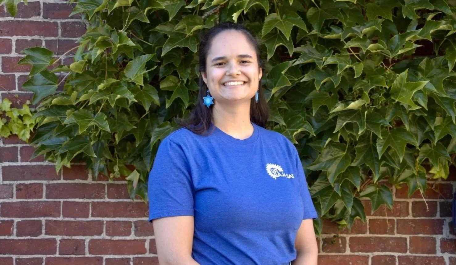 A woman with dark hair, wearing blue earrings and a blue T-shirt, standing outdoors in front of a brick wall and green leafy plant, smiling.