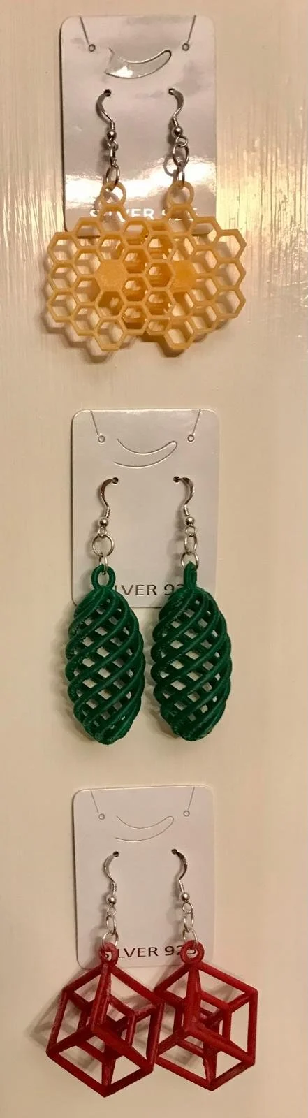 Three pairs of earrings hanging on a display card. The top pair are yellow honeycomb patterns, the middle pair are green lattice structures, and the bottom pair are red 3D cube shapes.