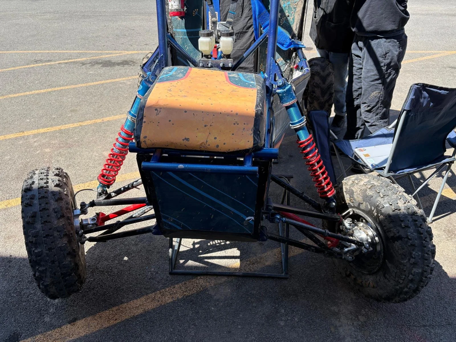 An off-road racing vehicle with large tires, red shocks, and a blue frame, displayed outdoors. People are standing around it, and a folding chair and a laptop are visible nearby.