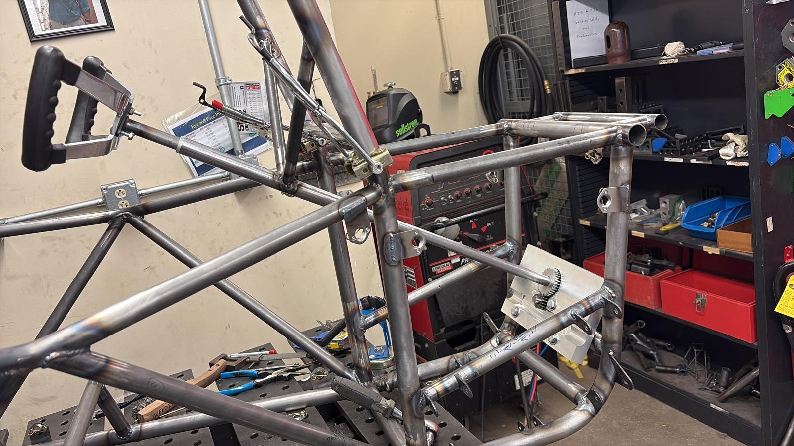 Metal framework of a race car in a workshop, with tools on the workbench and shelves in the background.