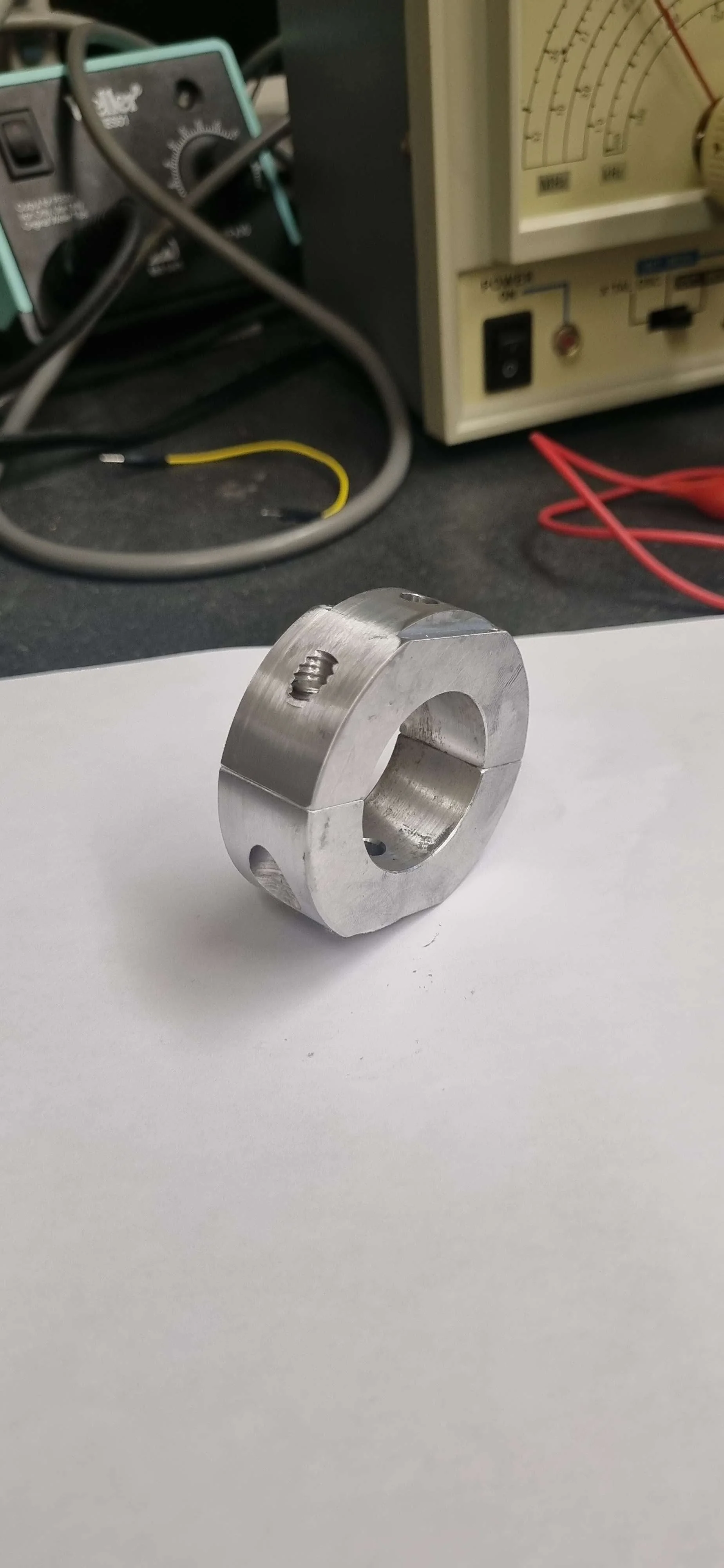 Metal mechanical component with a circular shape, threaded hole, and slots, placed on a white surface in a laboratory or workshop environment.