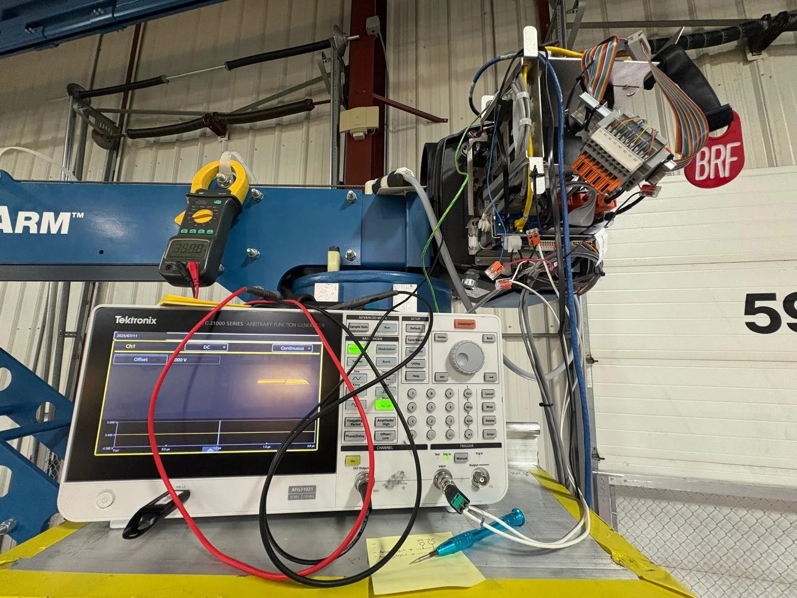 Electrical testing equipment setup on a blue work platform with various wires and tools, including a Tektronix generator and a multimeter, in an industrial or warehouse setting.