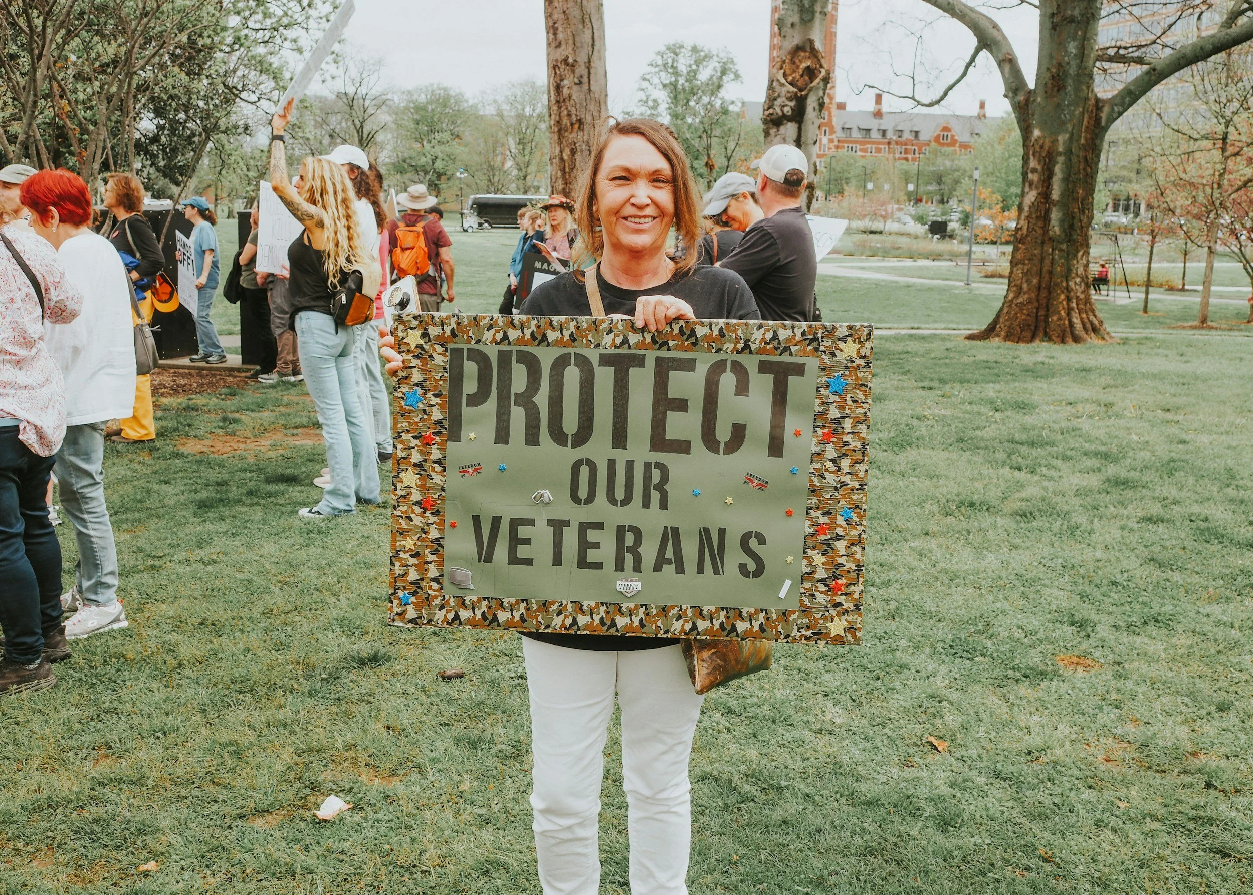 A woman smiling and holding a protest sign that reads 'Protect Our Veterans' at an outdoor rally, with other protesters and trees in the background.