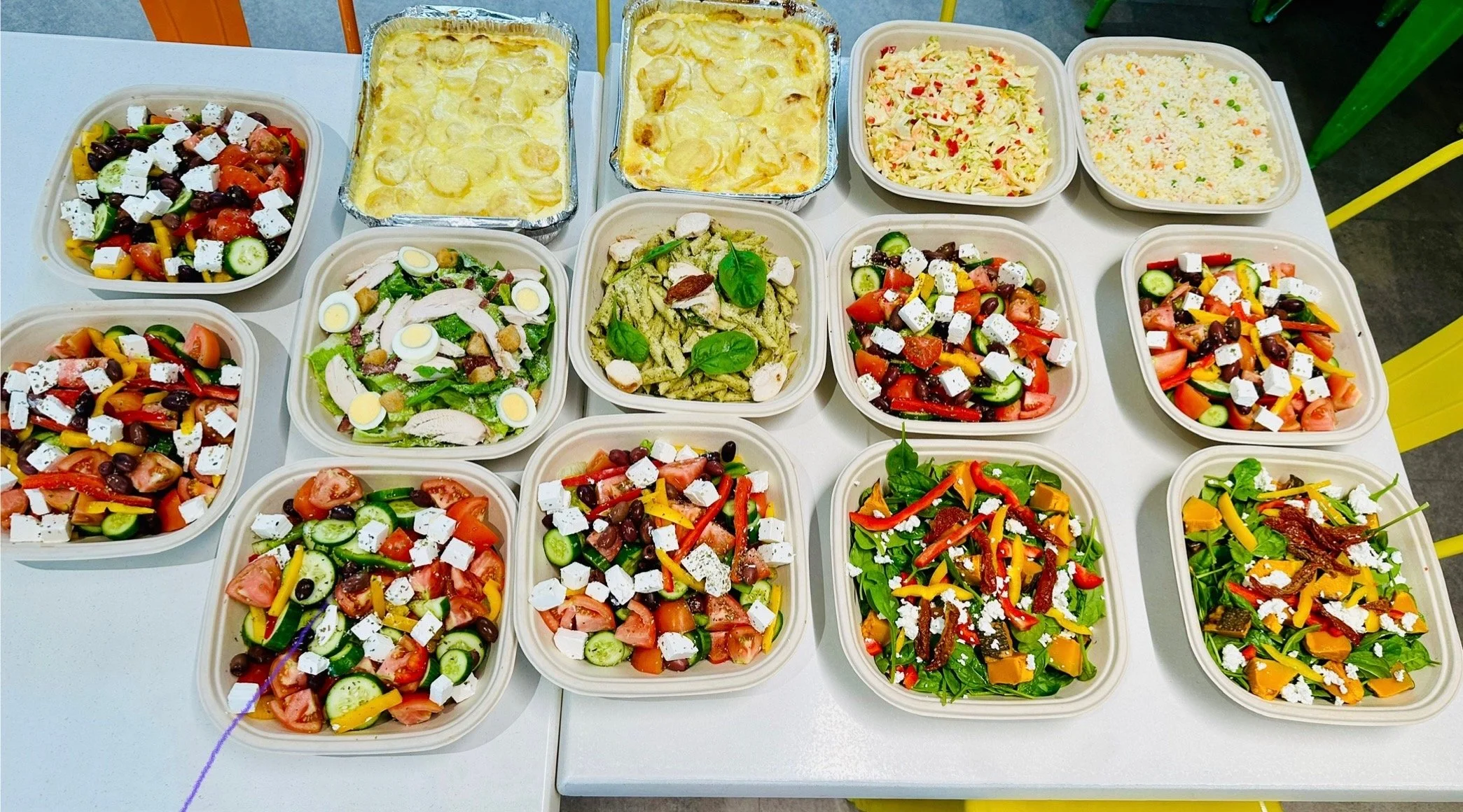 Assorted salads and side dishes on a white table, including pasta salad, vegetable salads with feta cheese, potato casserole, and rice.