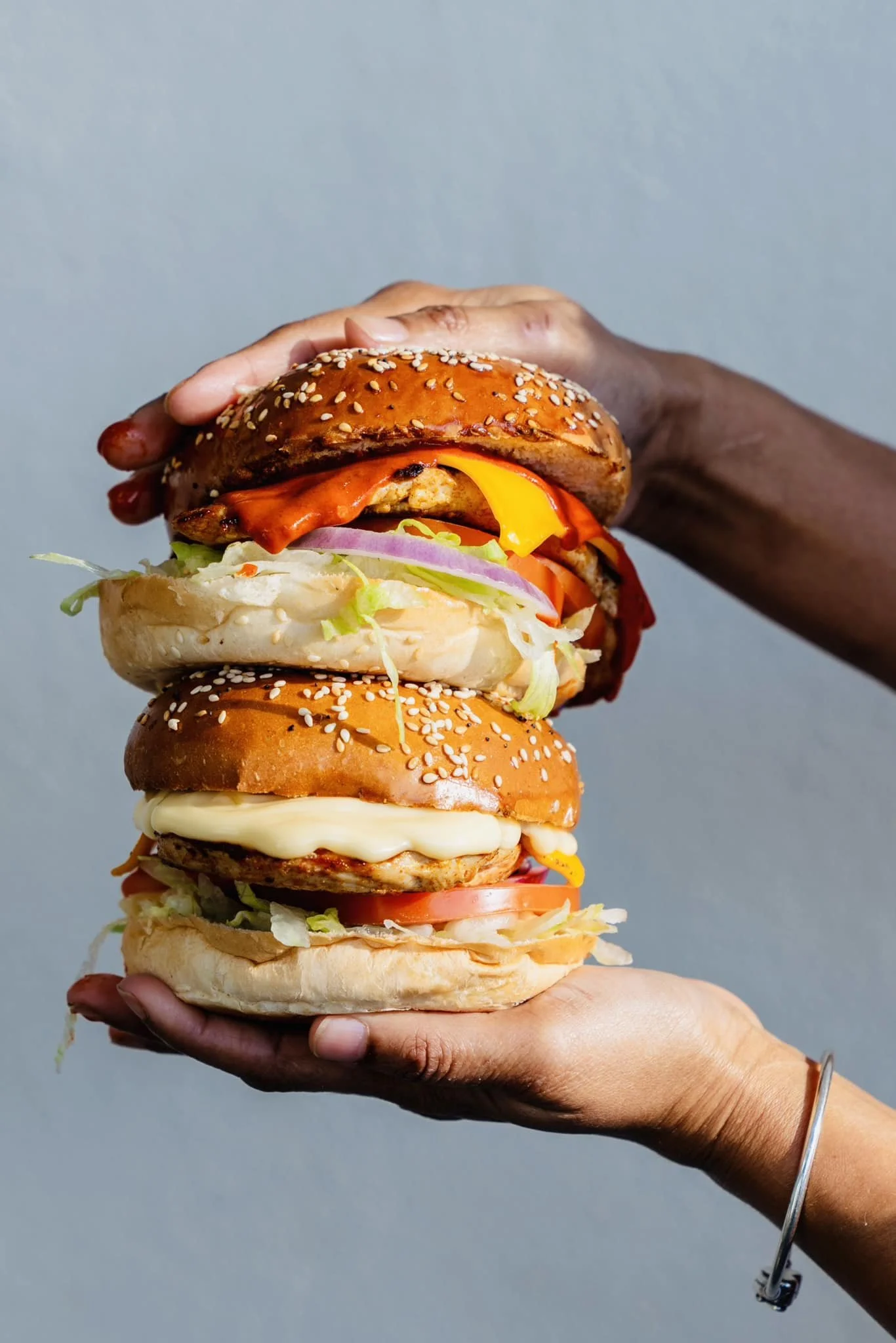 Person holding a large stack of three cheeseburgers with lettuce, tomato, onion, cheese, and sesame seed buns.