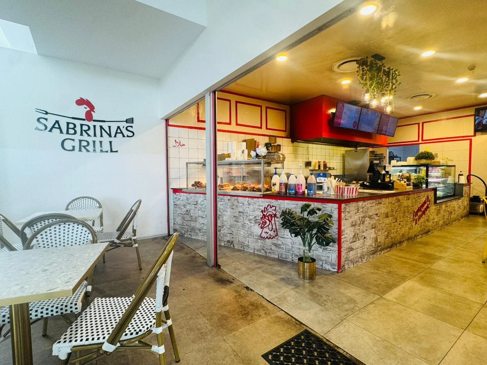 Interior view of Sabrina's Grill with a seating area on the left and a counter on the right. The seating includes round tables and chairs with black and white dotted pattern. The counter displays bottles and food items with digital screens above. The