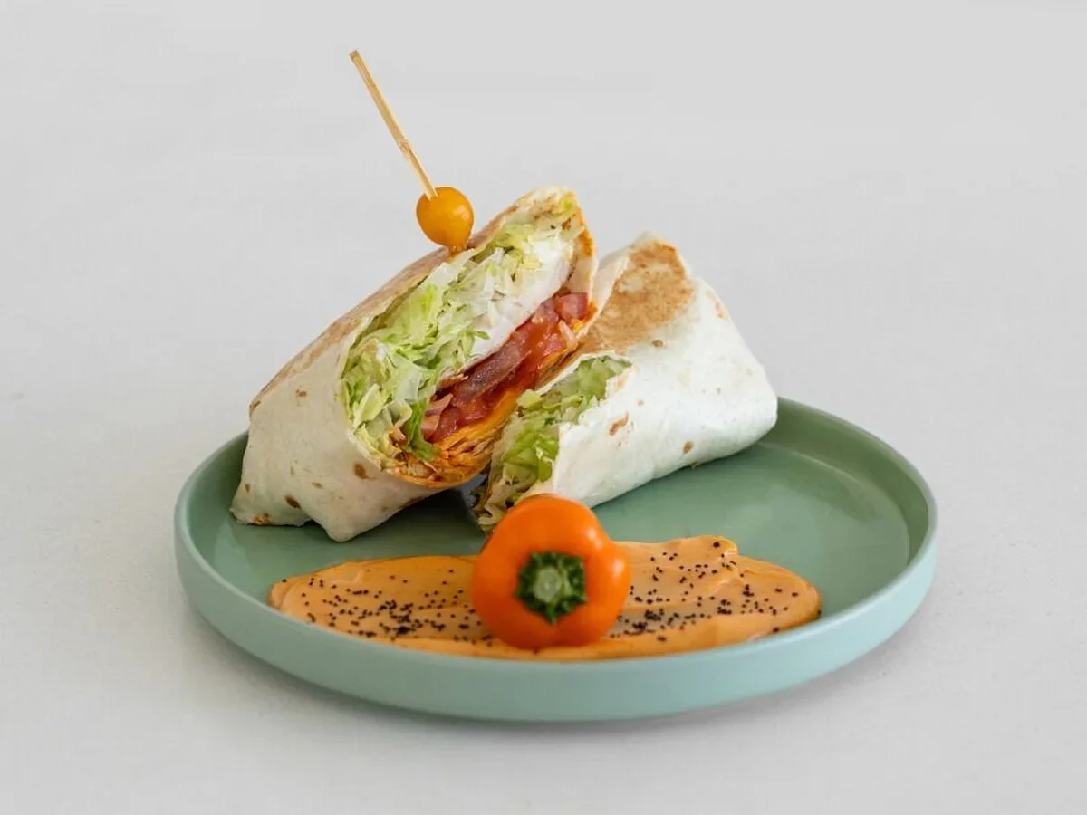 A wrap sandwich filled with lettuce, tomato, and bacon, garnished with a small yellow pepper and a special sauce.