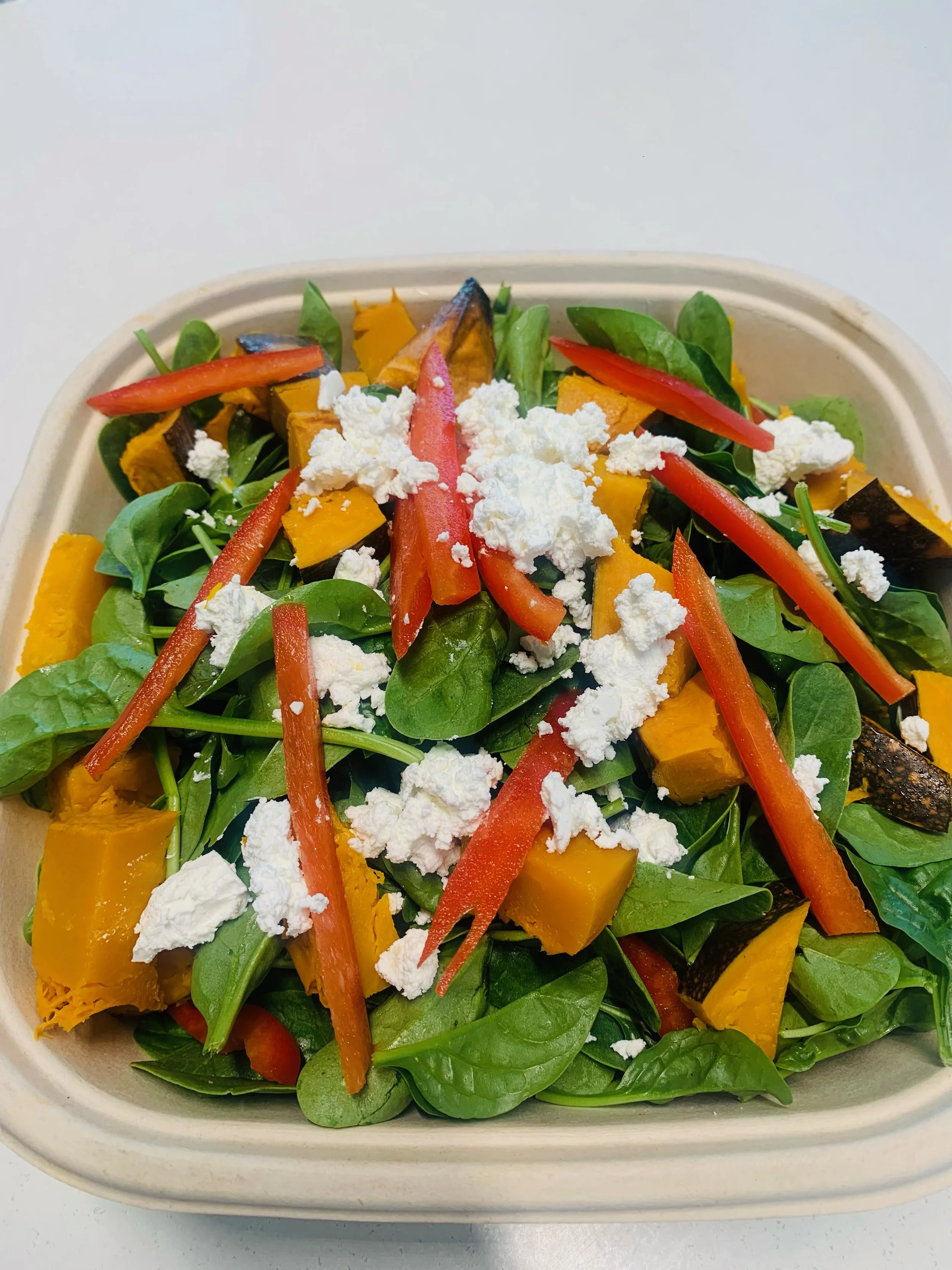 Fresh vegetable salad with spinach, roasted pumpkin, red peppers, and crumbled cheese in a biodegradable container.