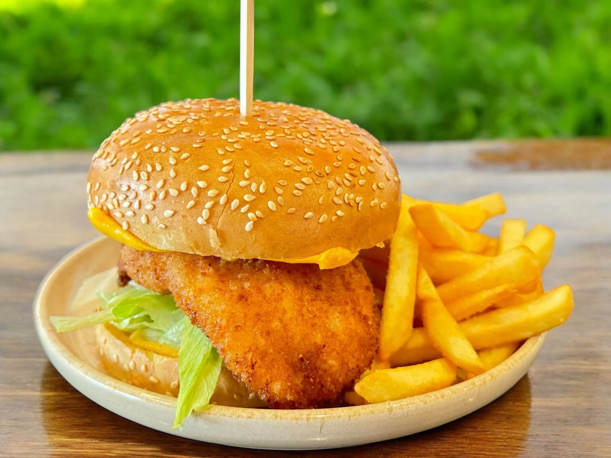 Burger with fried chicken fillet, lettuce, and cheese on a sesame seed bun, served with French fries on a beige plate.