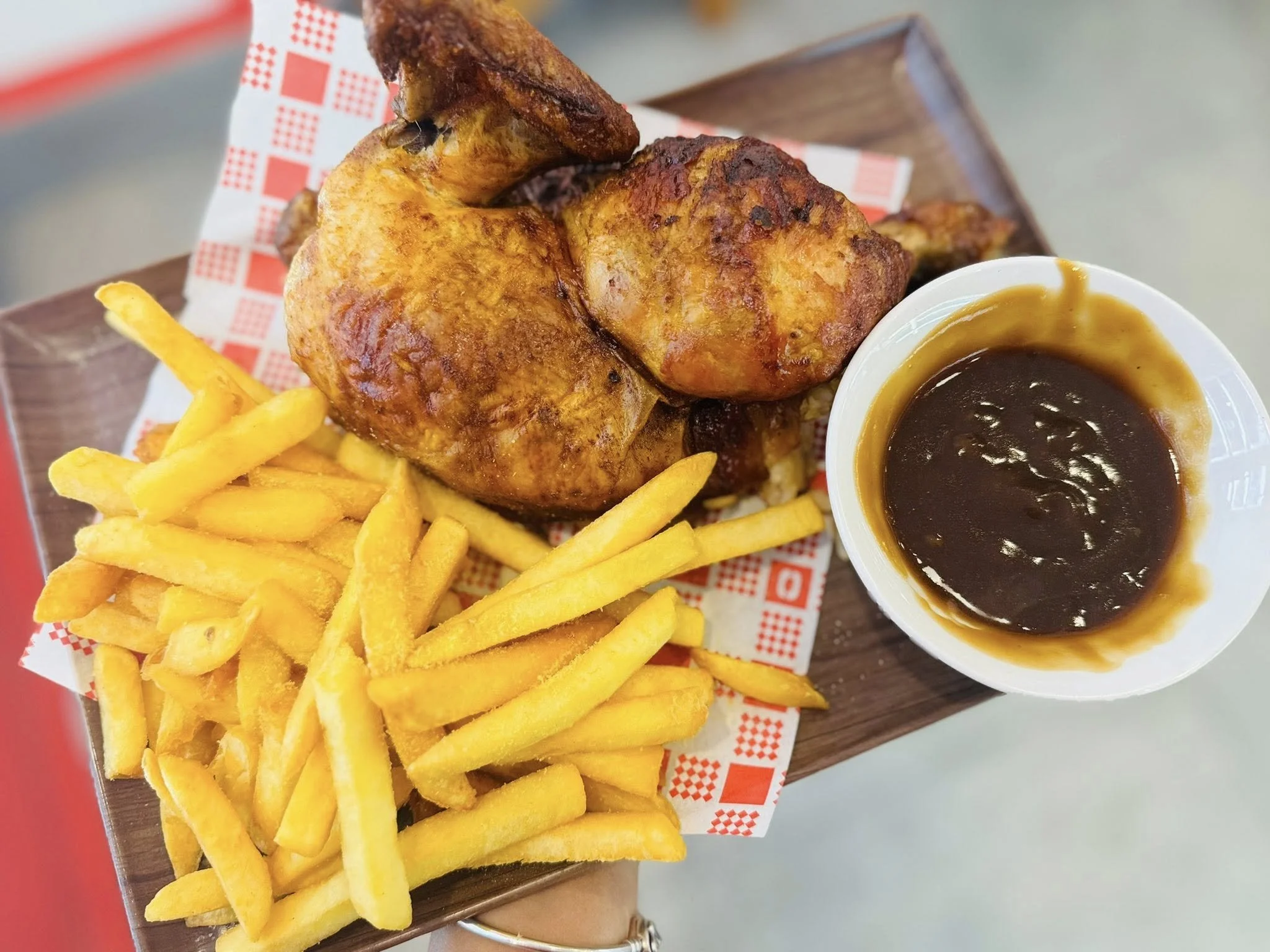 Fried chicken leg, French fries, and a cup of brown gravy on a wooden tray with red checkered paper.