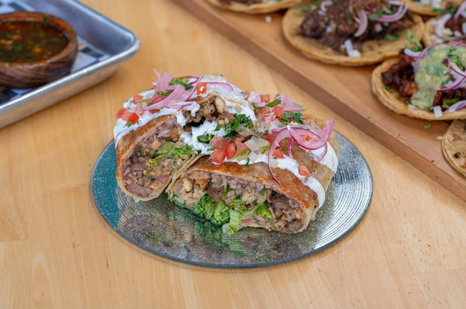 Burrito mexicano partido por la mitad, con ingredientes visibles como carne, verduras, crema, y salsa, servido en un plato de cerámica sobre una mesa de madera. En el fondo, una bandeja con otros tacos.
