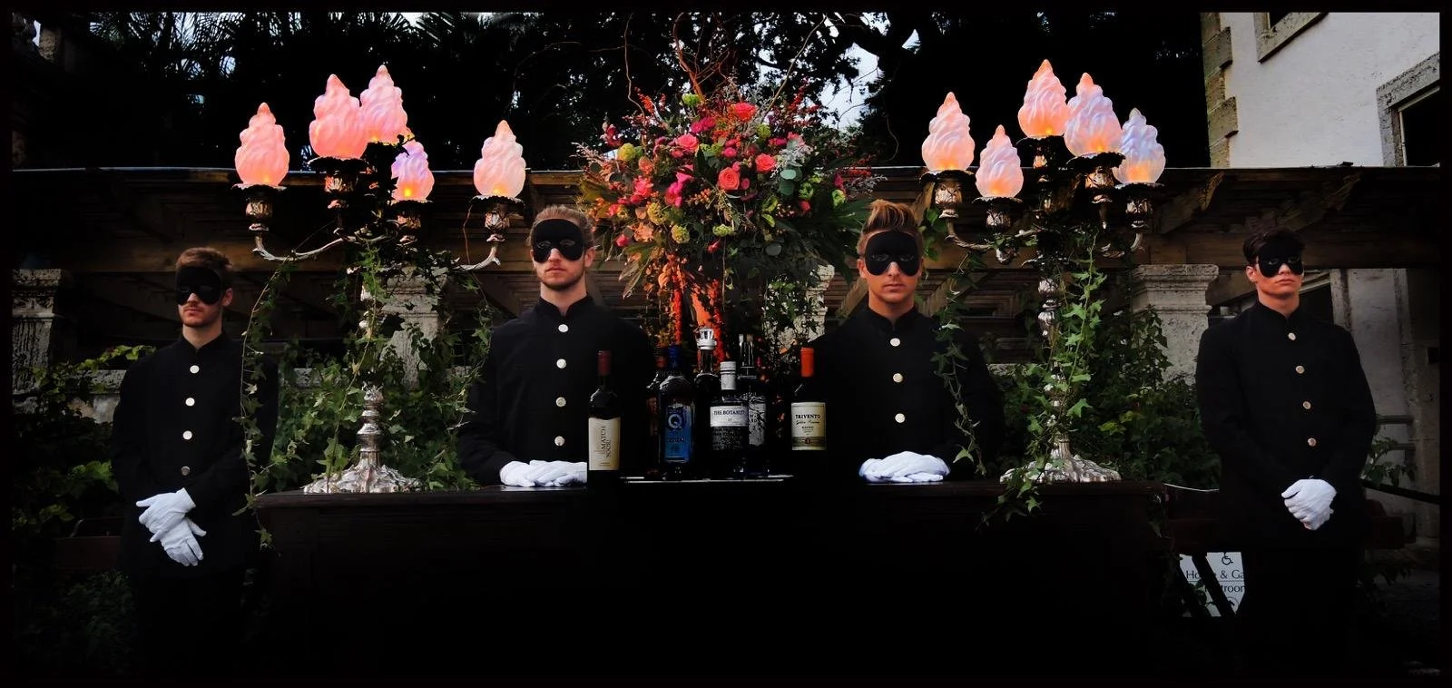 Four individuals dressed in black uniform with white gloves and eye masks stand behind a decorated table with bottles of alcohol and floral arrangements; they are outdoors at an event with decorative lighting and foliage.