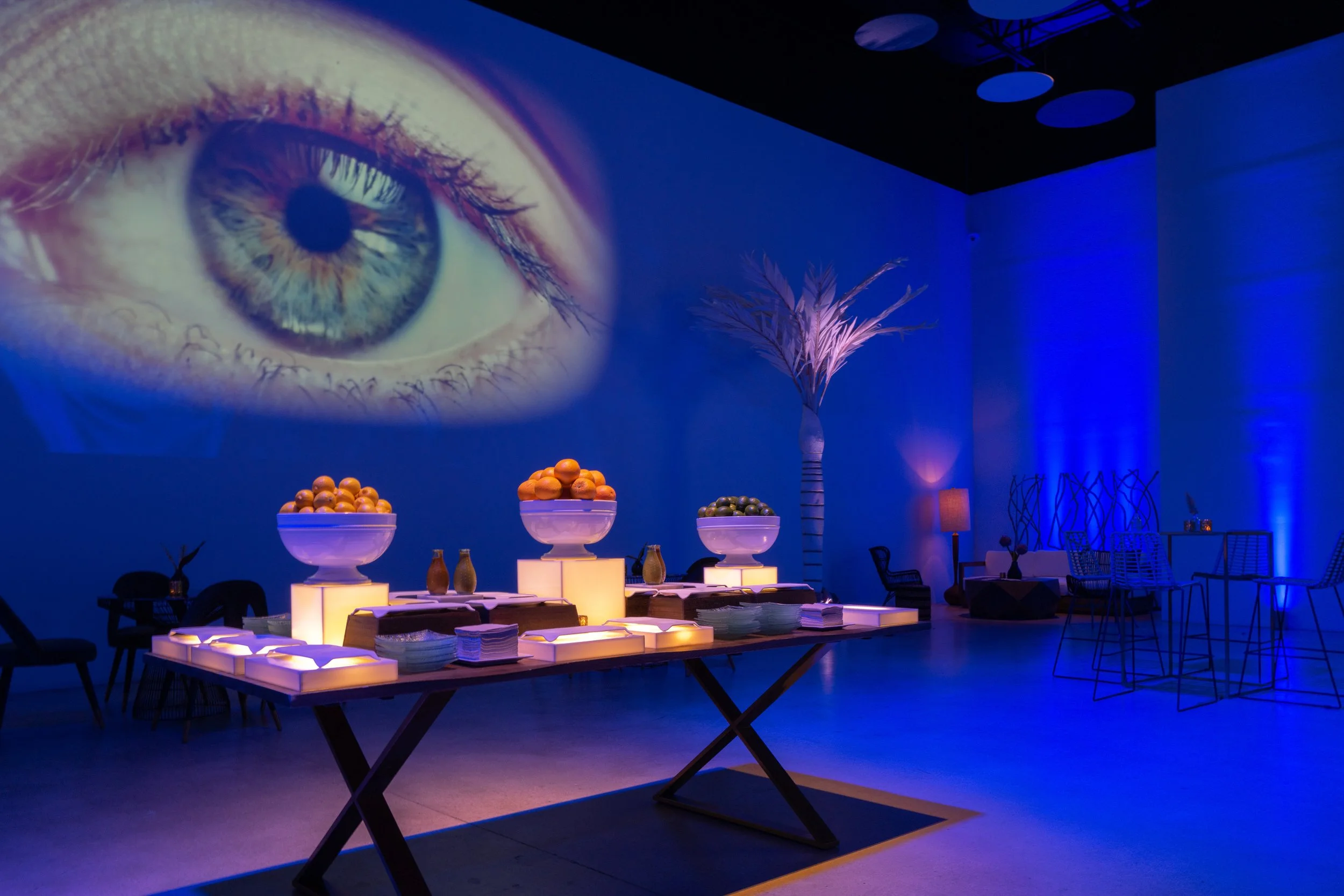 An indoor space with dim lighting and a large projected image of a human eye on a blue-lit wall. There are tables with lit cube bases, bowls of fruit, and chairs arranged around. Decorative vases and plants are also visible.