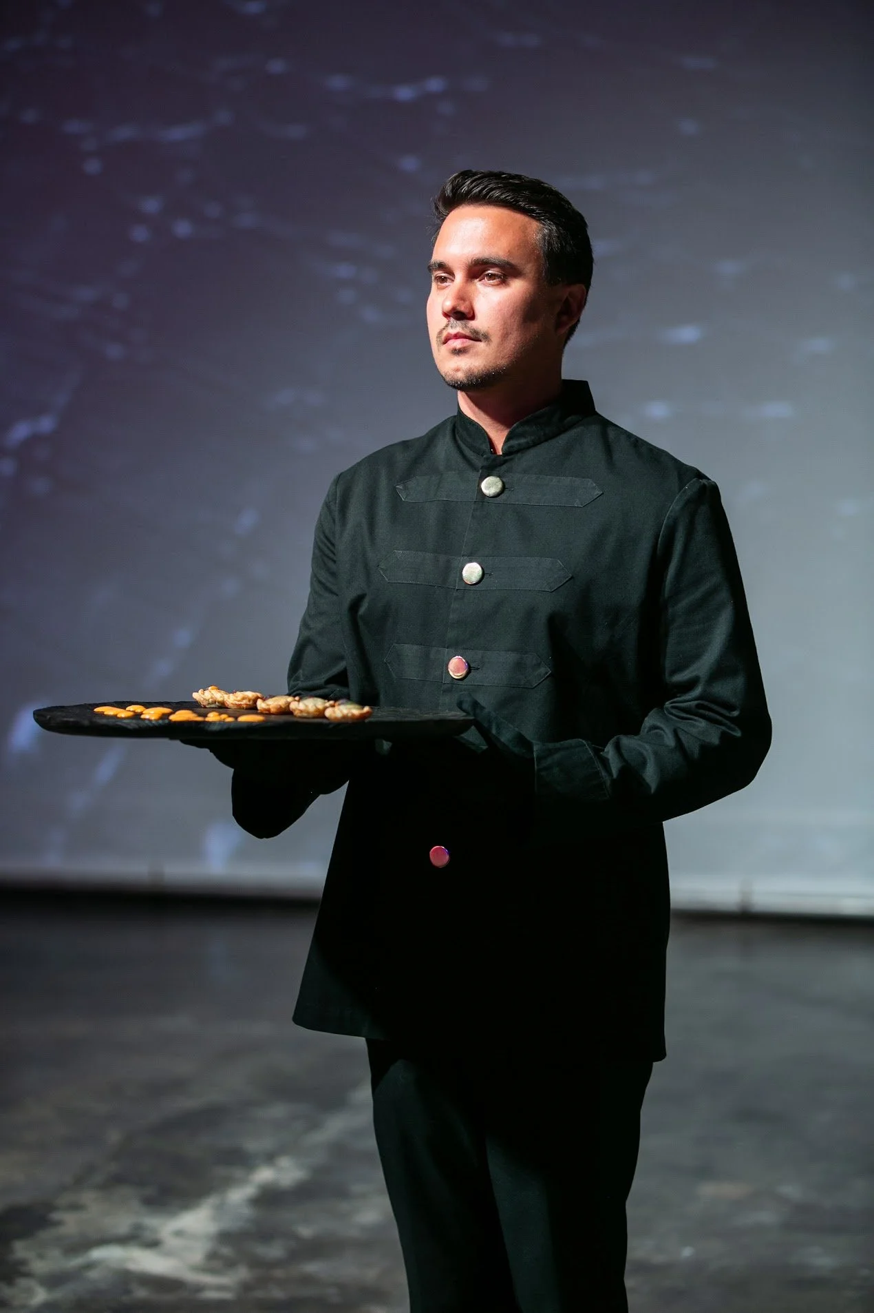 A man in a black chef's coat holding a black tray with food, standing on a dark stage with a blurry background.