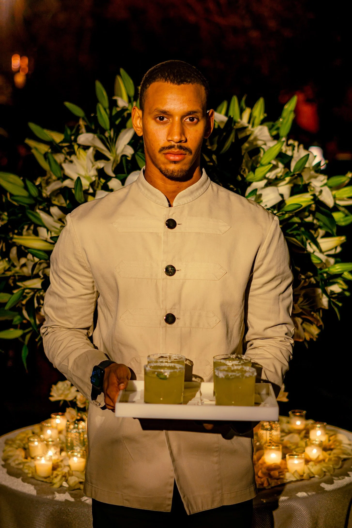 A man in a beige chef jacket holding a tray with four glasses of green cocktails at an elegant evening event with candles and floral decorations.
