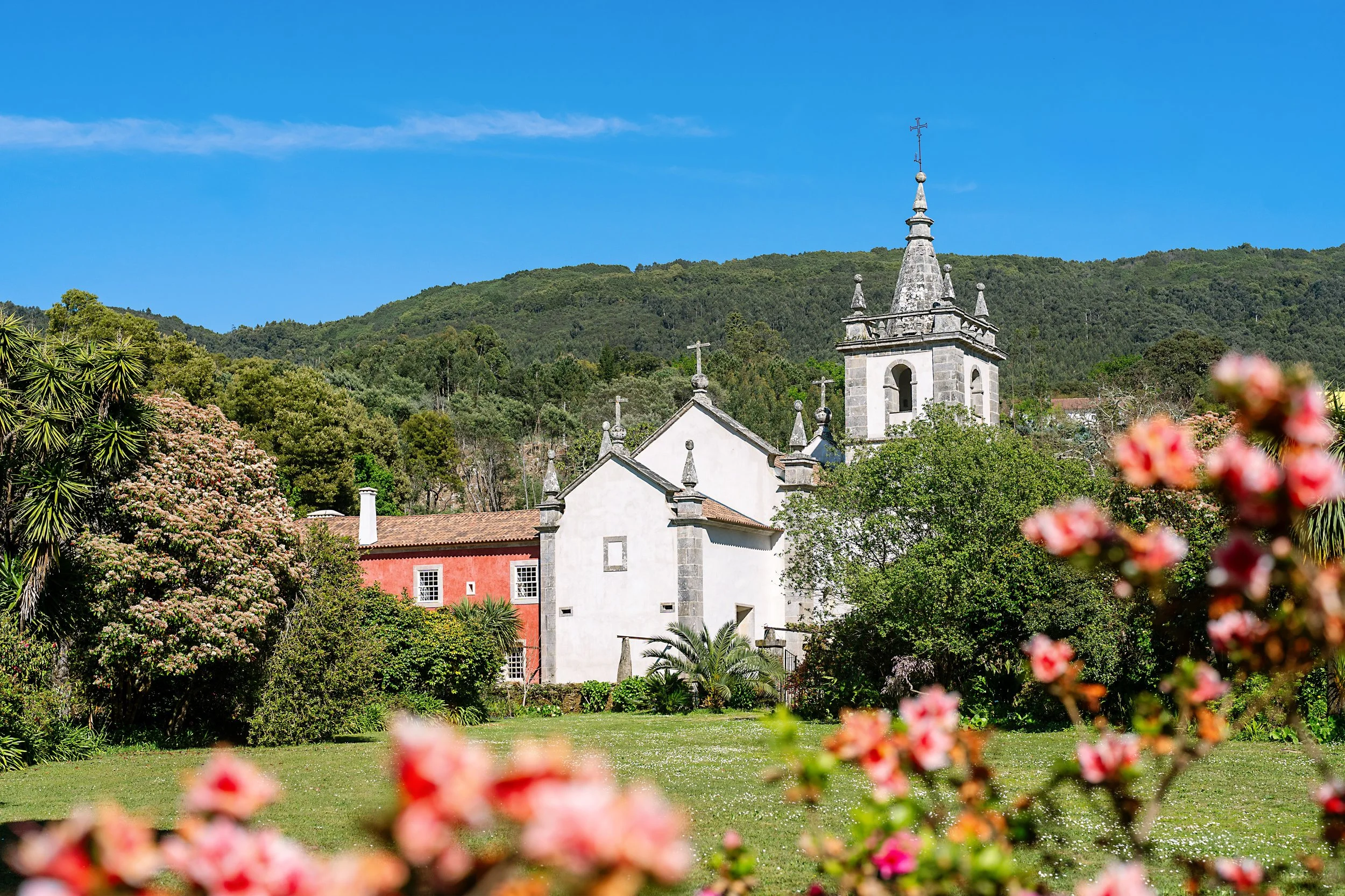 A scenic view of a church at the Convento de Cabanas.  A venue and hotel in Portugal.   Image is with a tall steeple topped by a cross, surrounded by lush greenery and colorful flowers in the foreground.