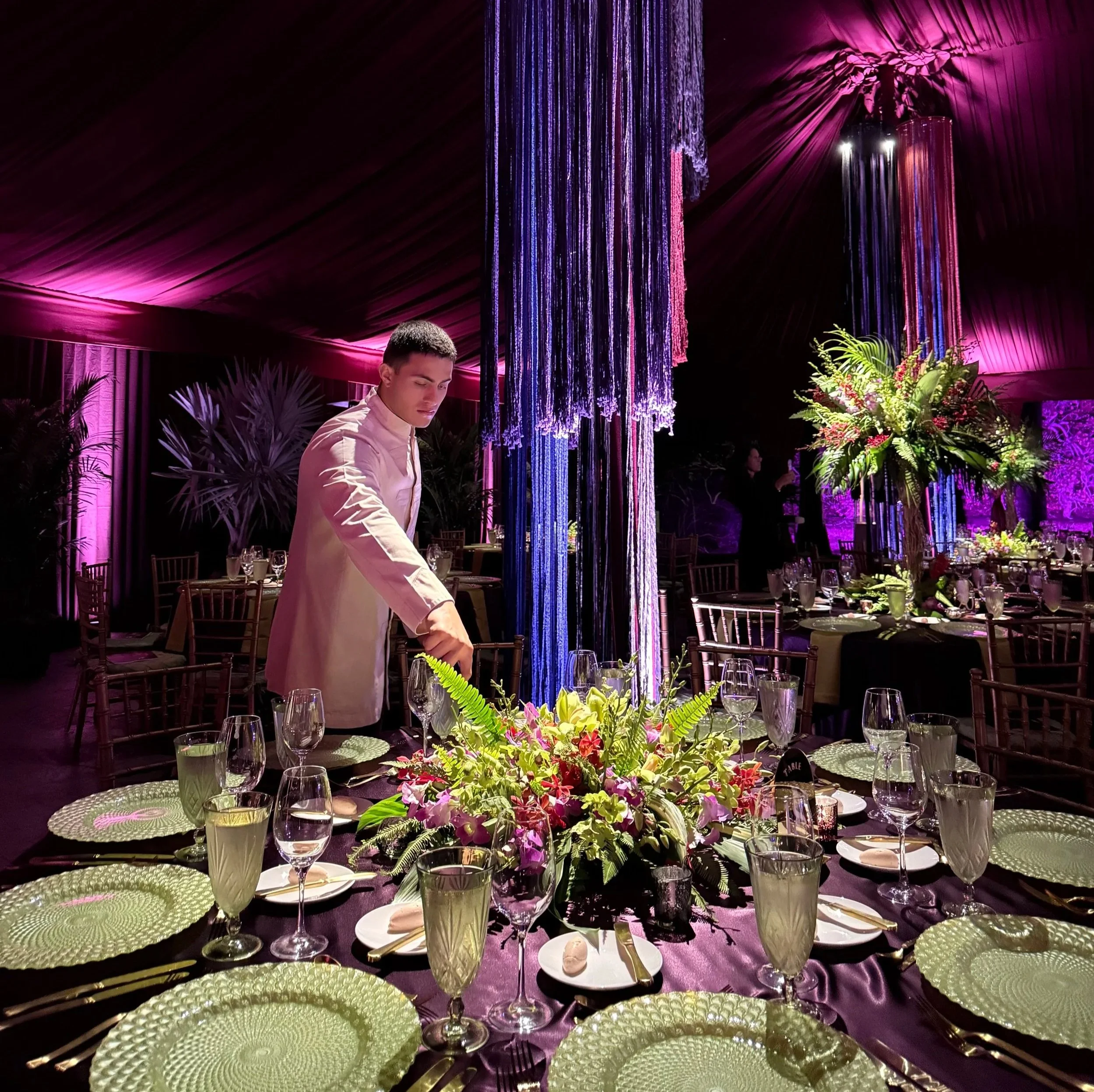 A young man in a light-colored tuxedo stands at a decorated banquet table with a colorful floral centerpiece, surrounded by wine glasses, plates, and green textured napkins, under purple lighting and floral decorations.