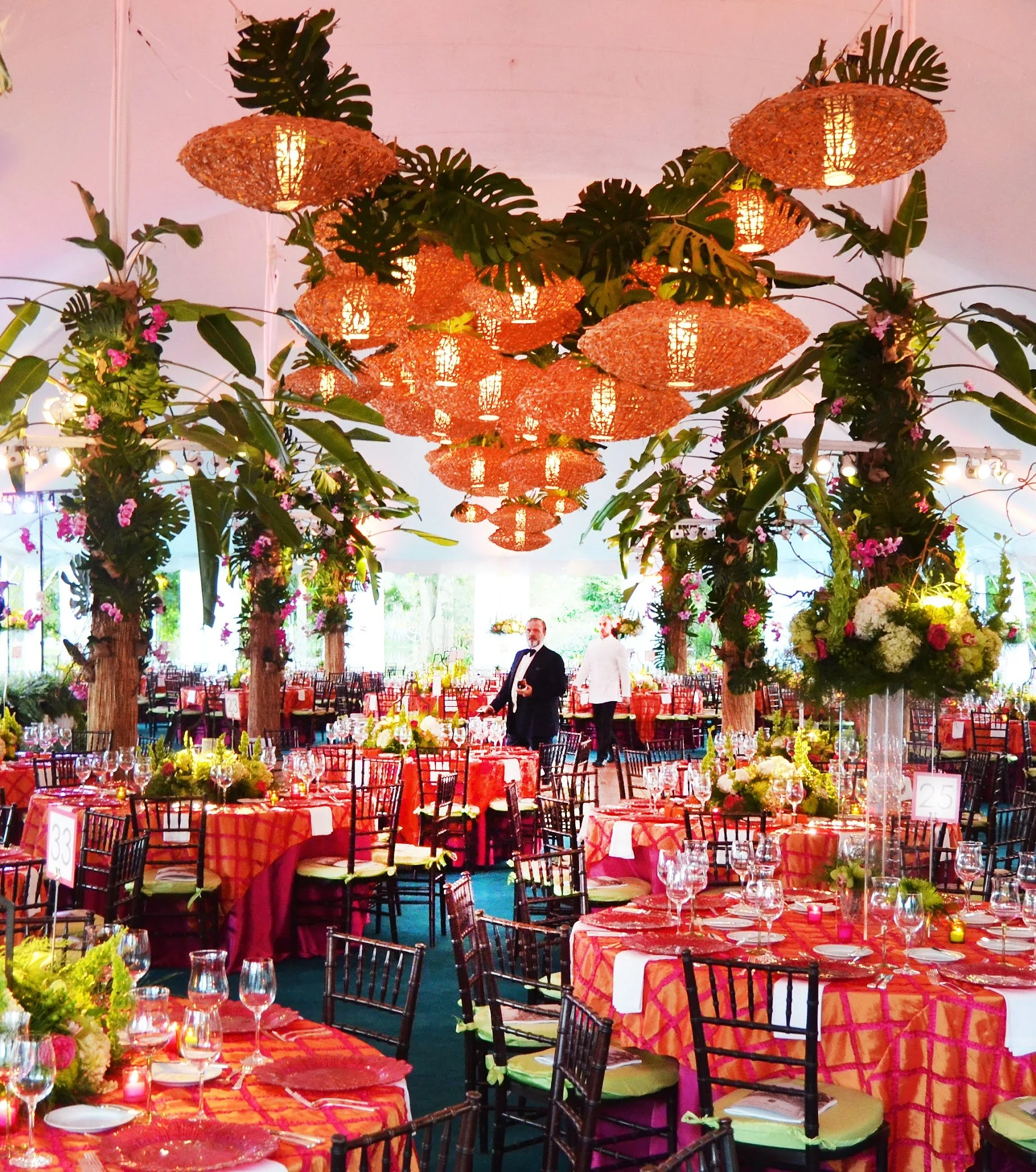 Decorated wedding reception area with round tables covered in red patterned tablecloths, elegant glassware, floral centerpieces, and lush green foliage and pink flowers hanging from wooden tree trunks. Large wicker and leaf-shaped light fixtures hang from the ceiling, illuminating the space with warm light.