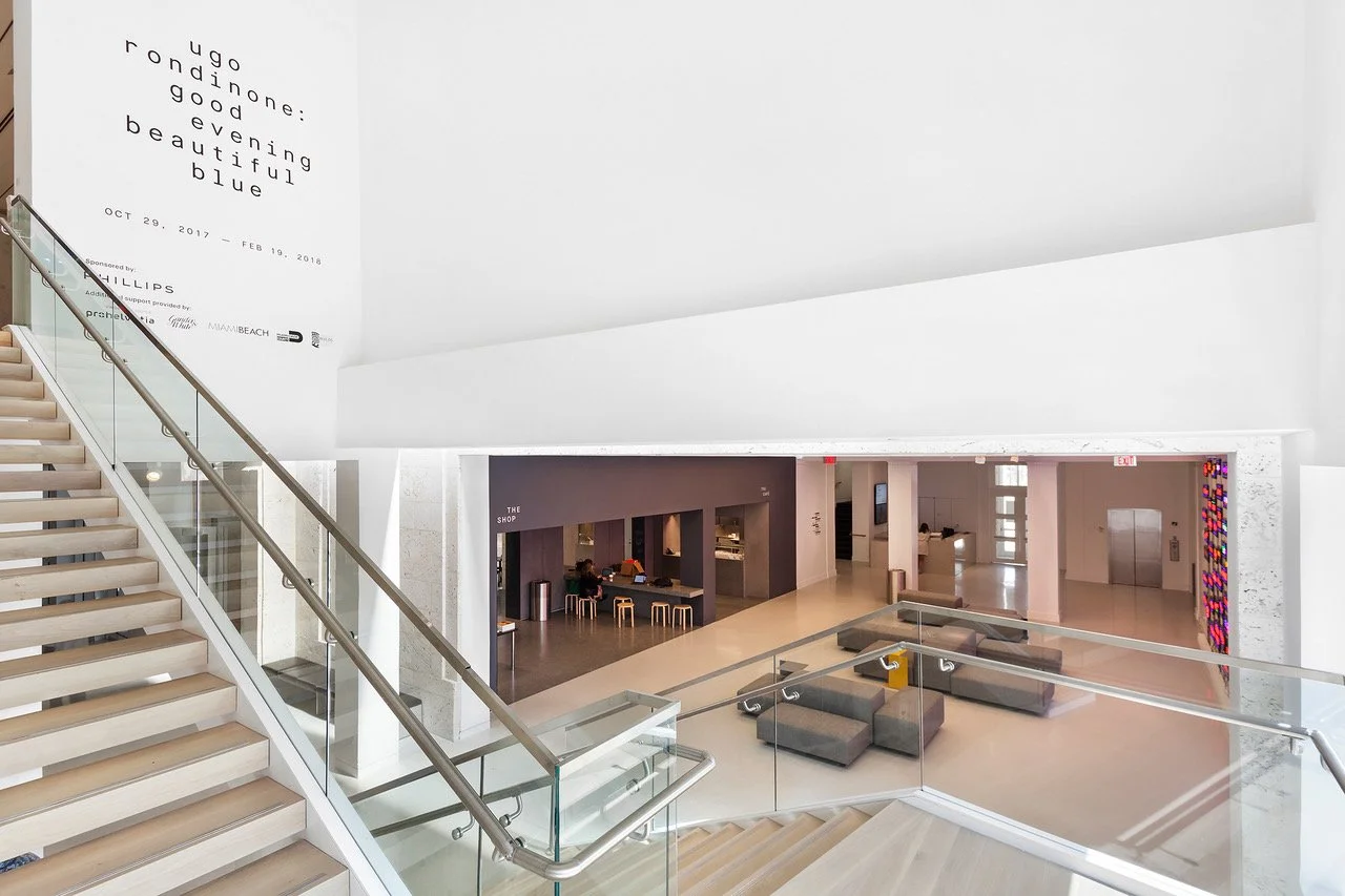 Interior view of a modern, bright building with stairs, a seating area with couches, and a shop with bar stools, featuring minimalist decor and natural lighting.