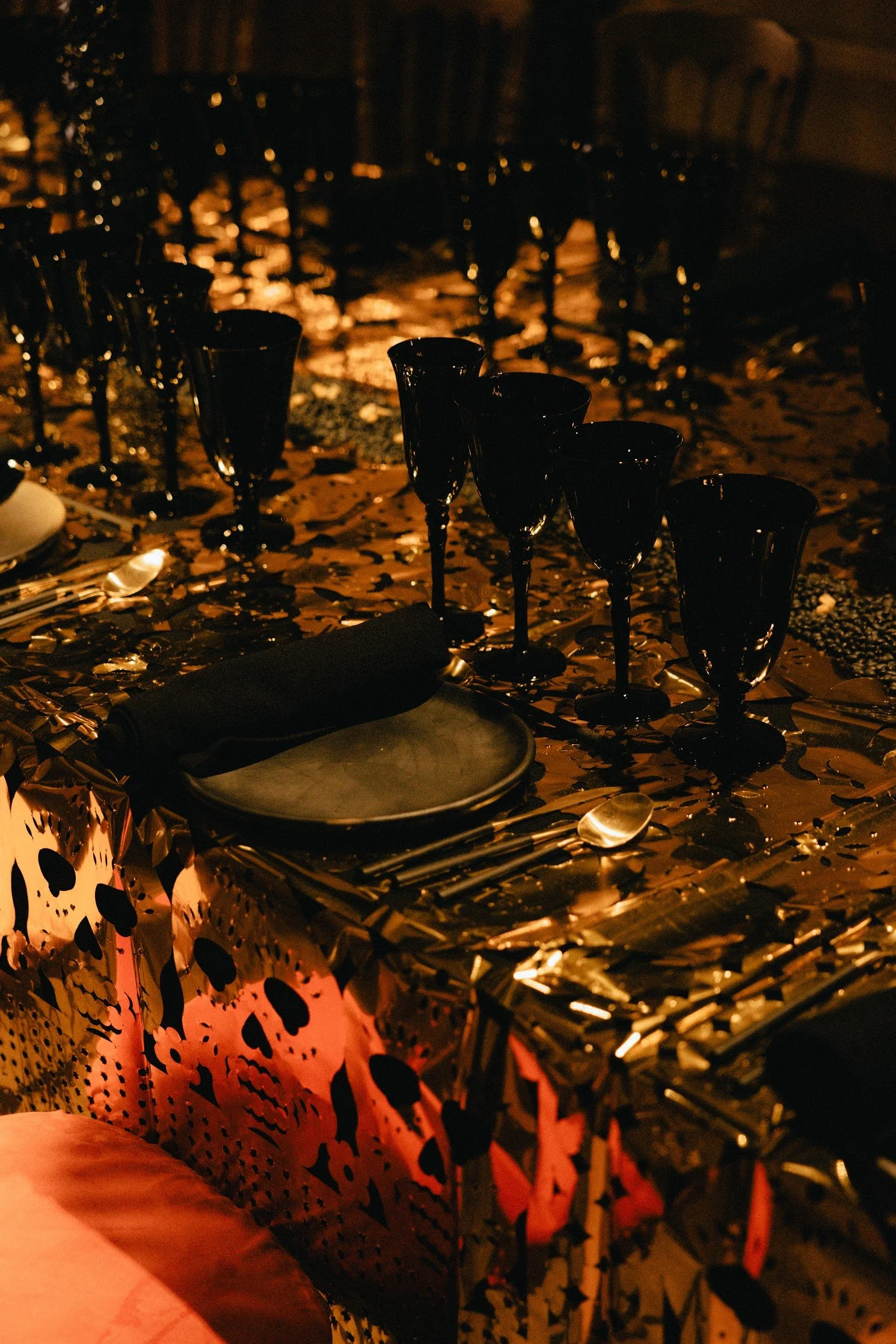 A table decorated for Halloween with black champagne glasses and a pumpkin-shaped tablecloth with black skulls. The table is covered with gold confetti, and there are silver utensils and black plates on it. The scene is dimly lit with a warm, orange glow.
