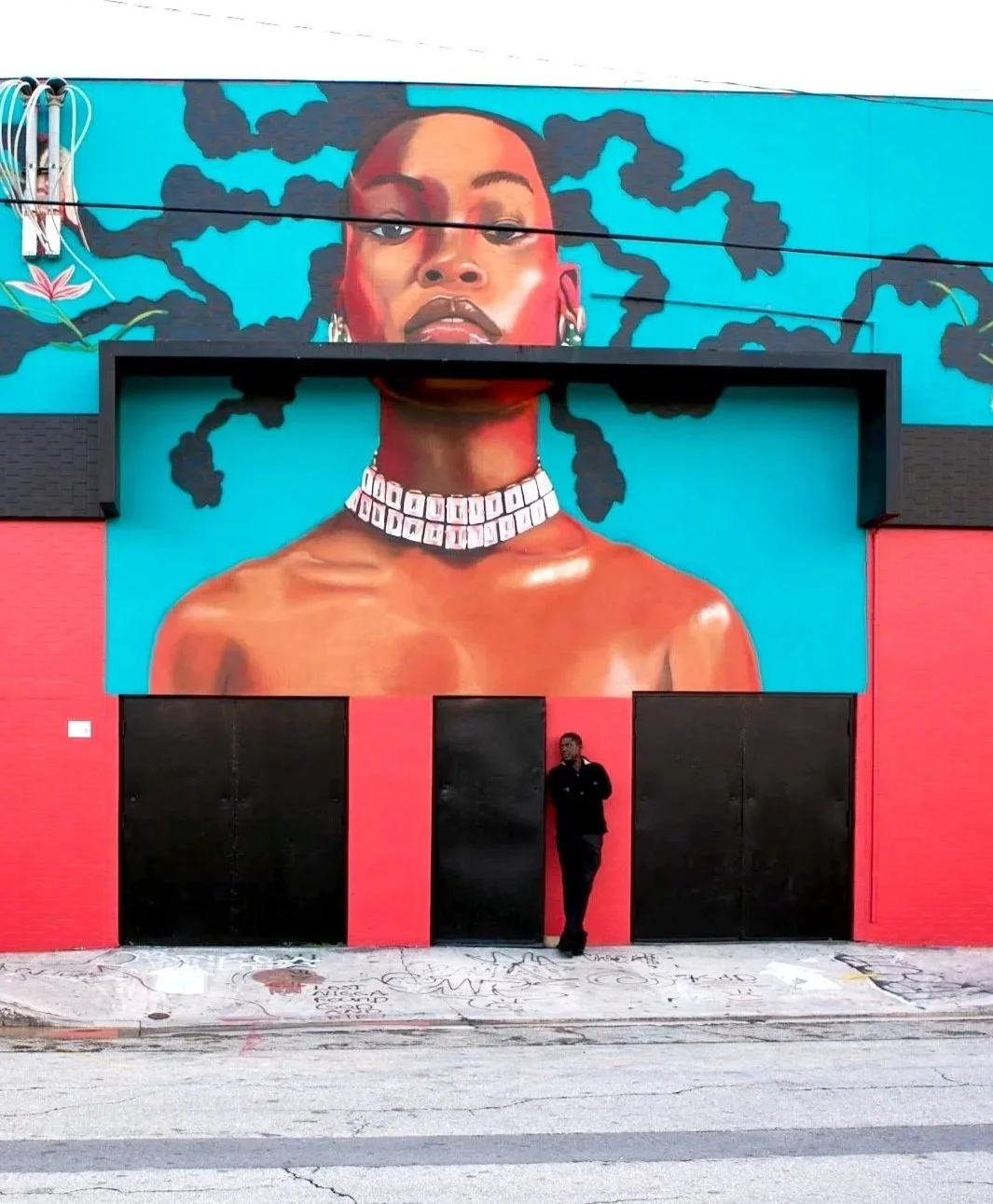 Colorful mural of a woman with voluminous black hair, wearing jewelry, displayed above a black and red building with a man standing in front.