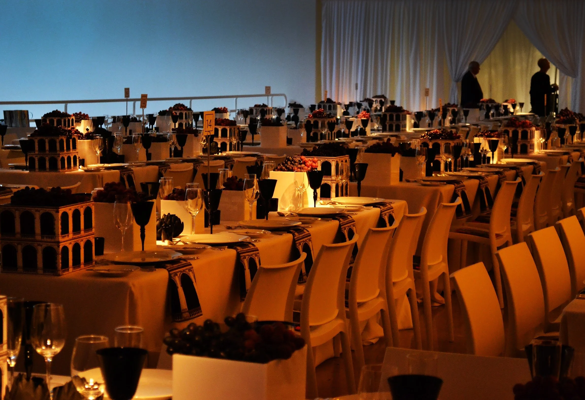 An elegant banquet hall with rows of tables decorated with model architecture and fruit displays, dim lighting, and two silhouetted individuals near a stage or curtained area.