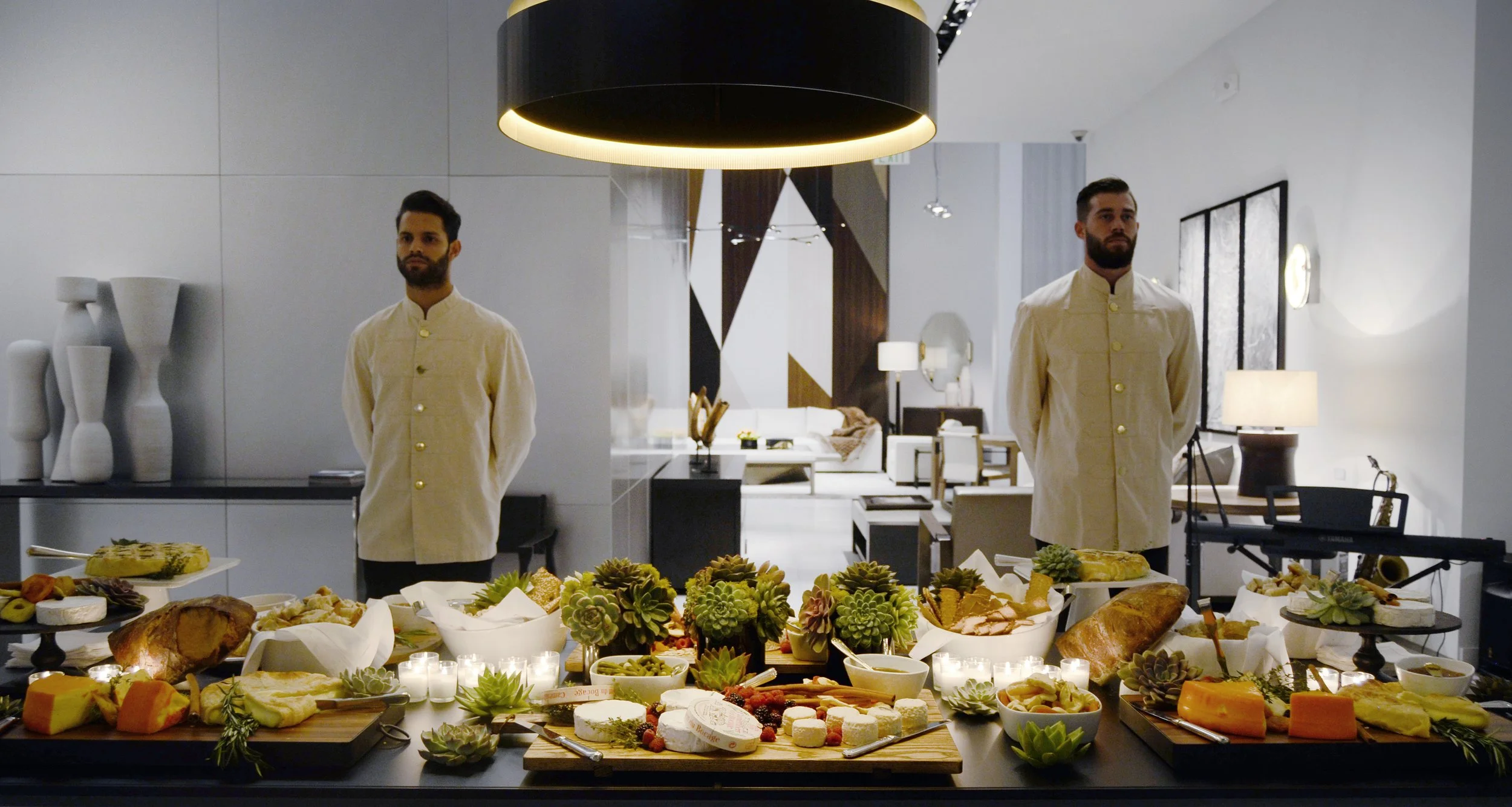 Two men in beige jackets standing behind a table filled with various foods and desserts in a modern, well-lit room.