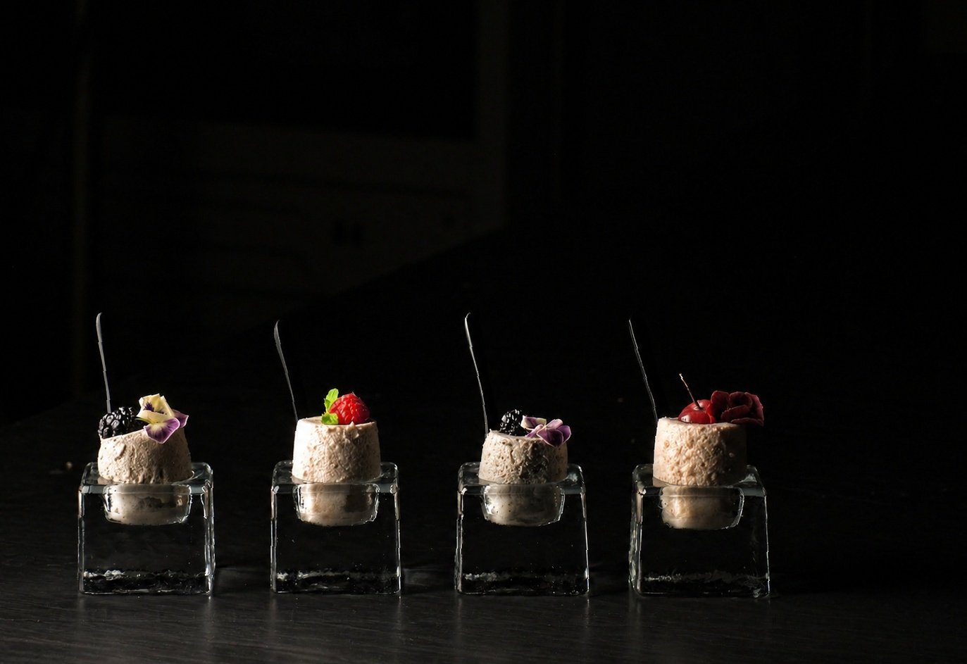 Four small desserts topped with berries and edible flowers, each placed on a clear glass stand against a dark background.