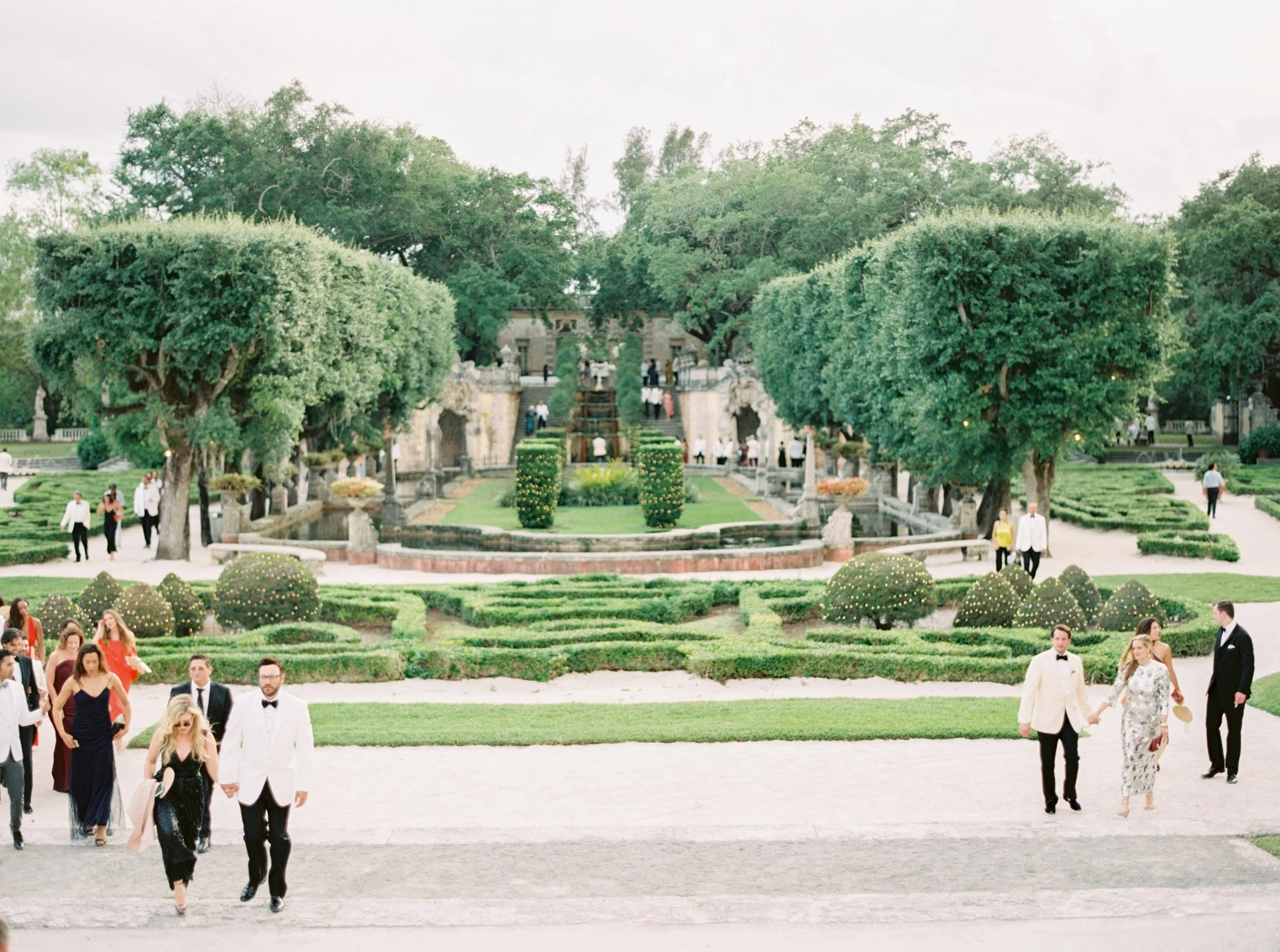People dressed in formal attire walking on a sandy path in a lush, well-manicured garden with large, trimmed trees and decorative shrubs, with a grand building in the background during a daytime outdoor event.