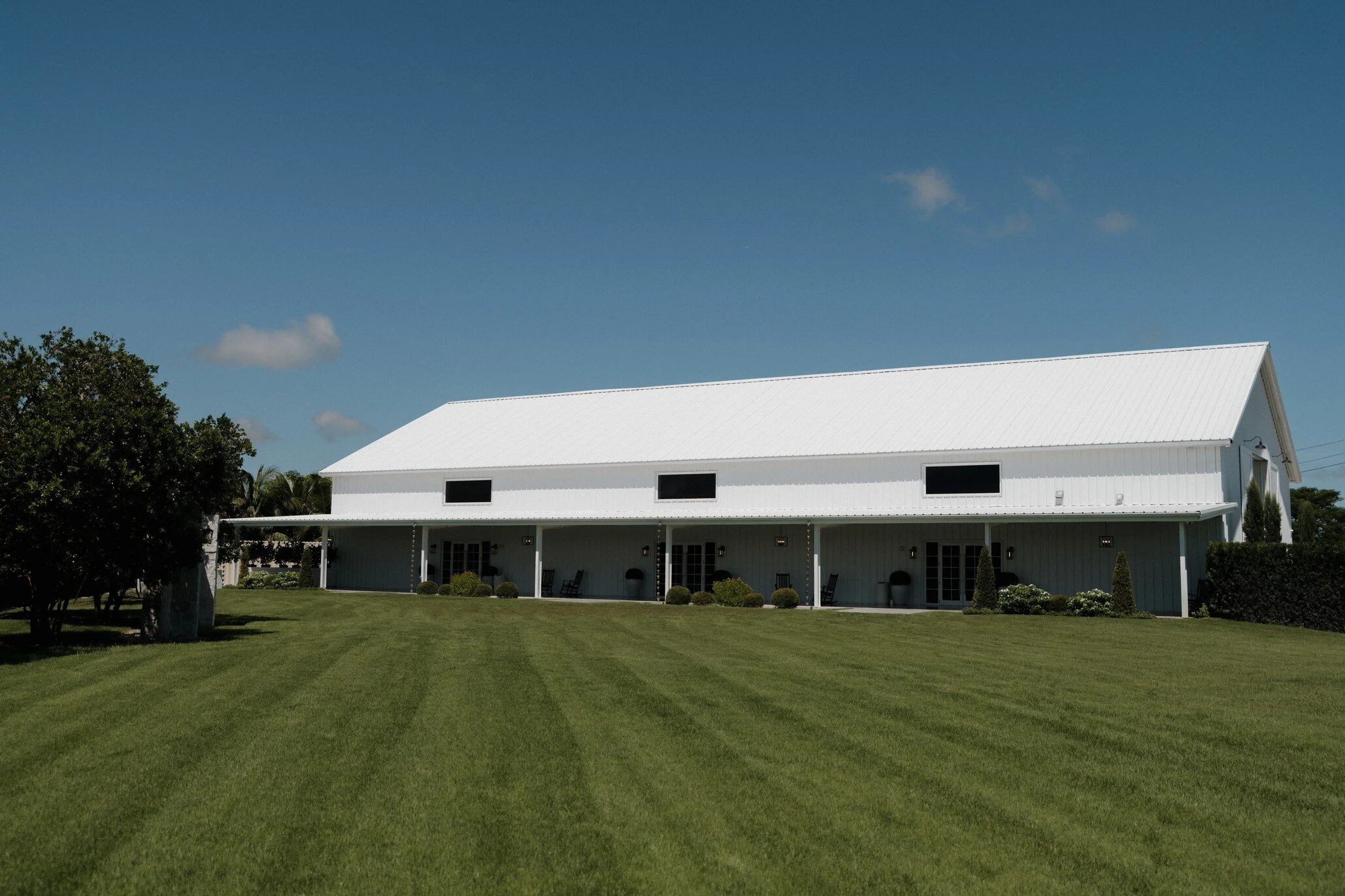 A white barn with a steep metal roof and a covered porch, surrounded by green lawn and trees, under a blue sky with a few clouds.