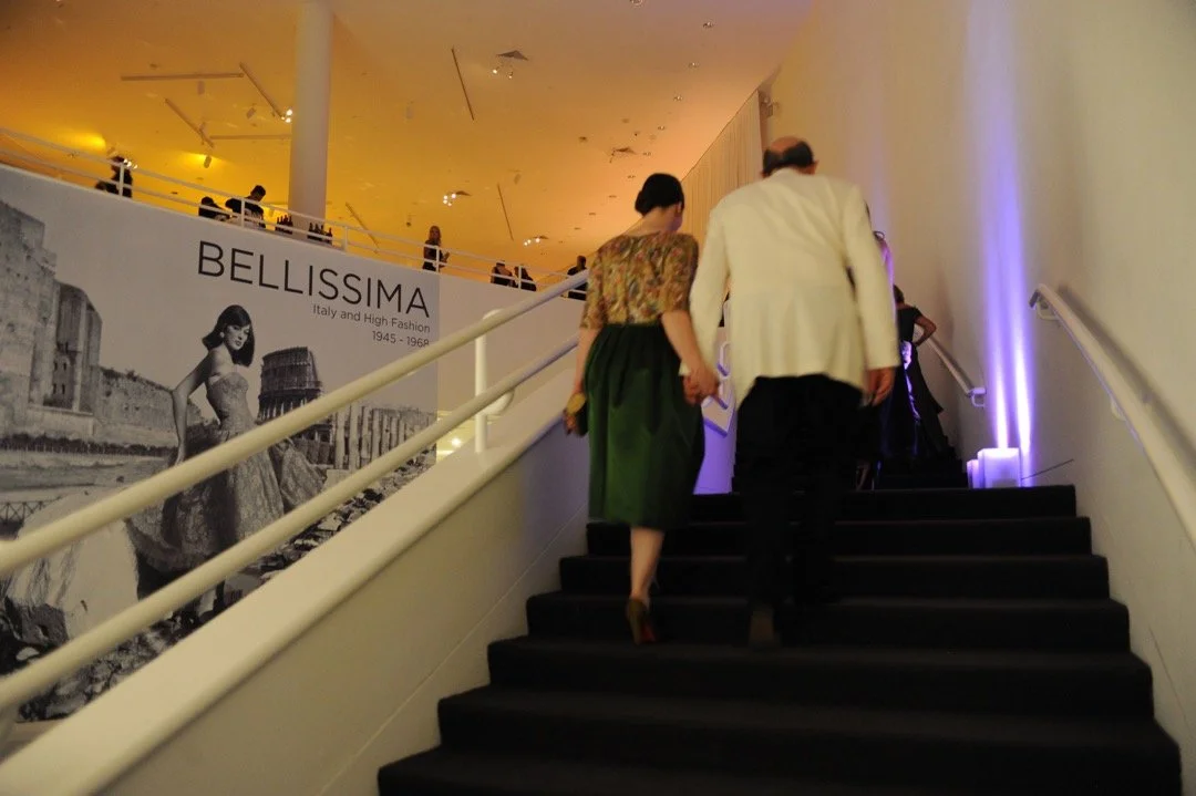 Two people ascending a staircase at an art exhibition, with a large black and white banner titled 'BELISSIMA' featuring a woman in a vintage dress and historic Greek architecture, illuminated with warm and purple lighting.