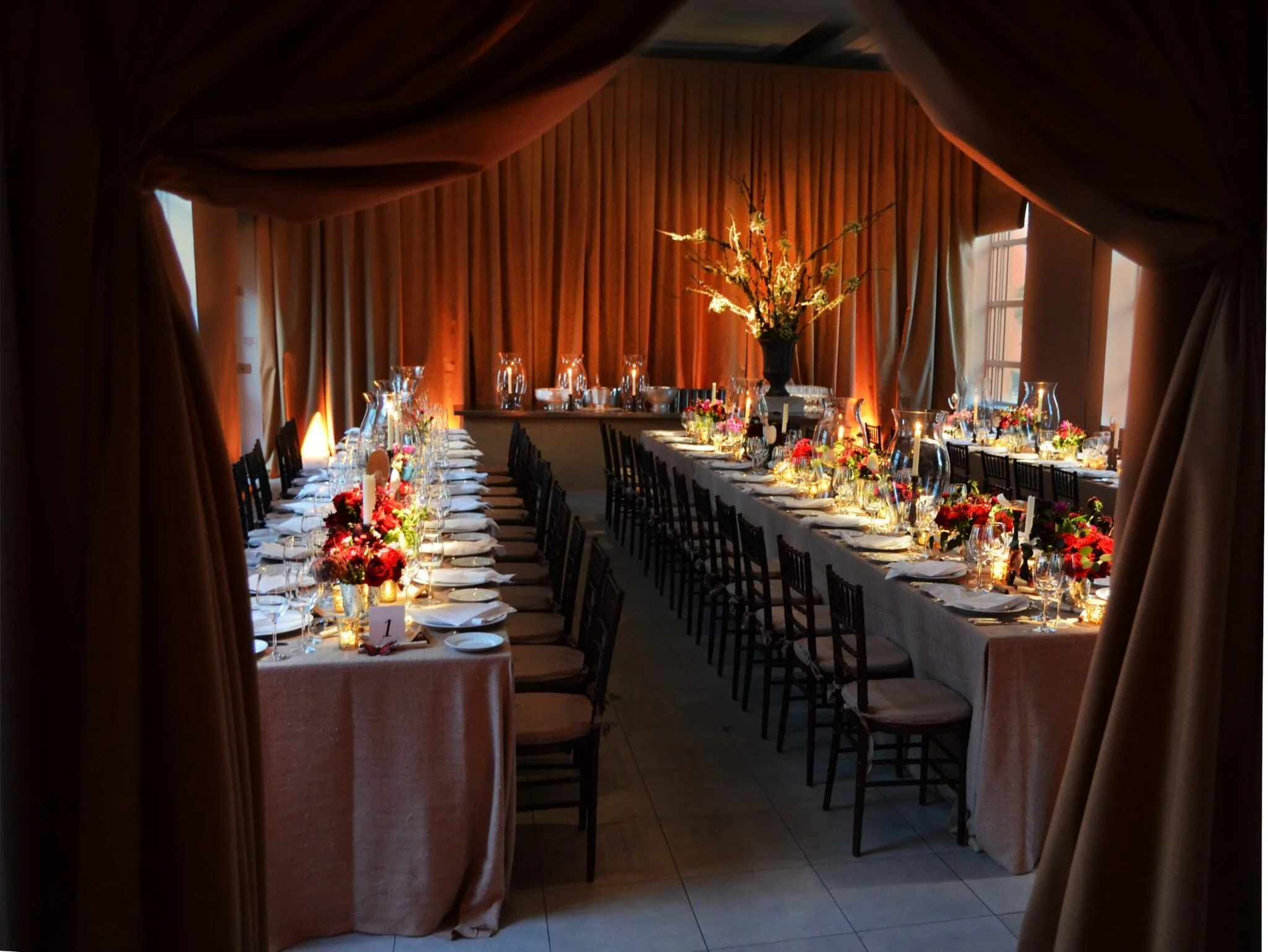 Elegant banquet dining room with long tables decorated with floral centerpieces, candles, and glassware, framed by heavy draped curtains.