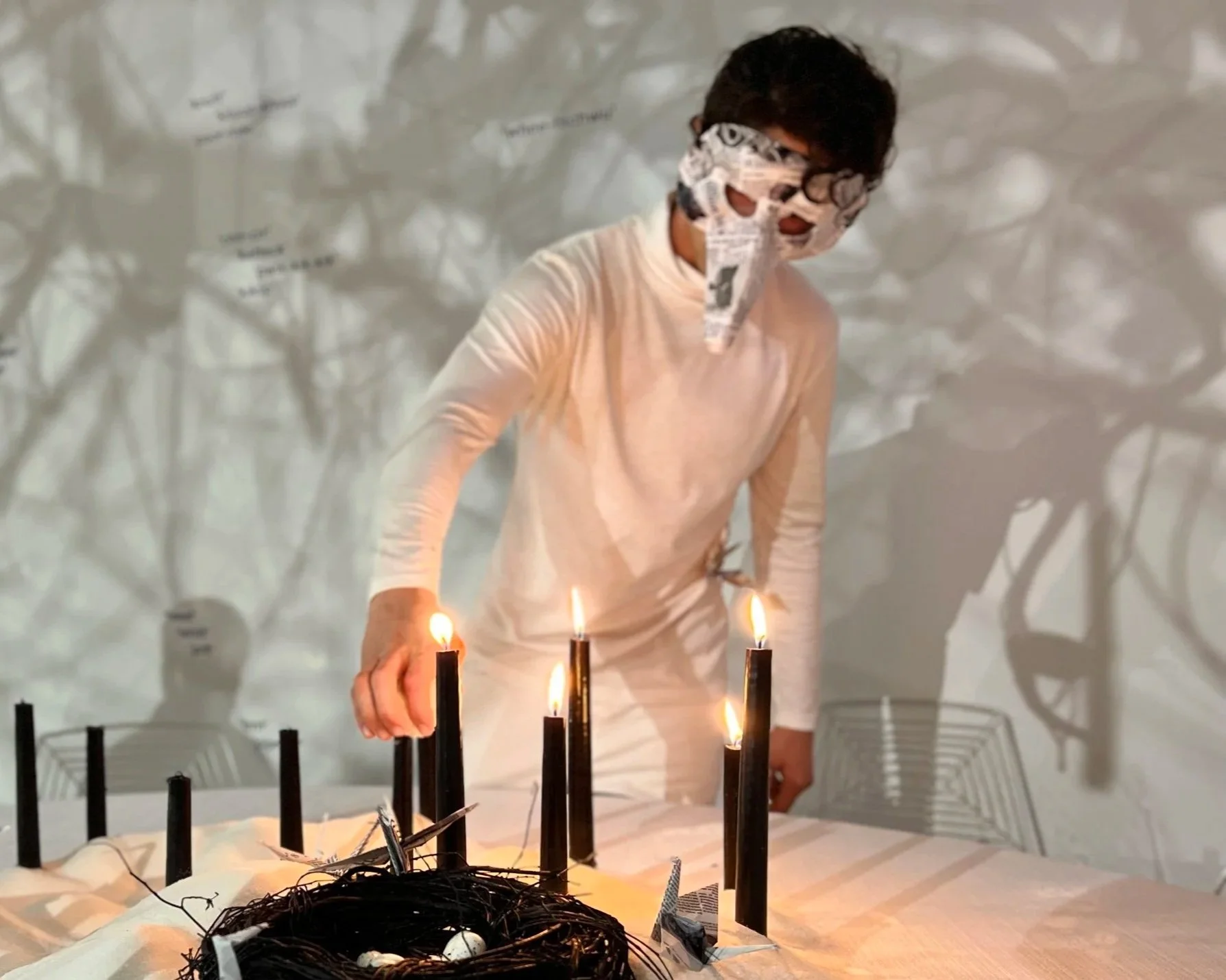 A person wearing a newspaper mask stands behind a table with lit candles arranged on it. The table is covered with newspaper and clouds of shadows and text are cast on the wall behind them.
