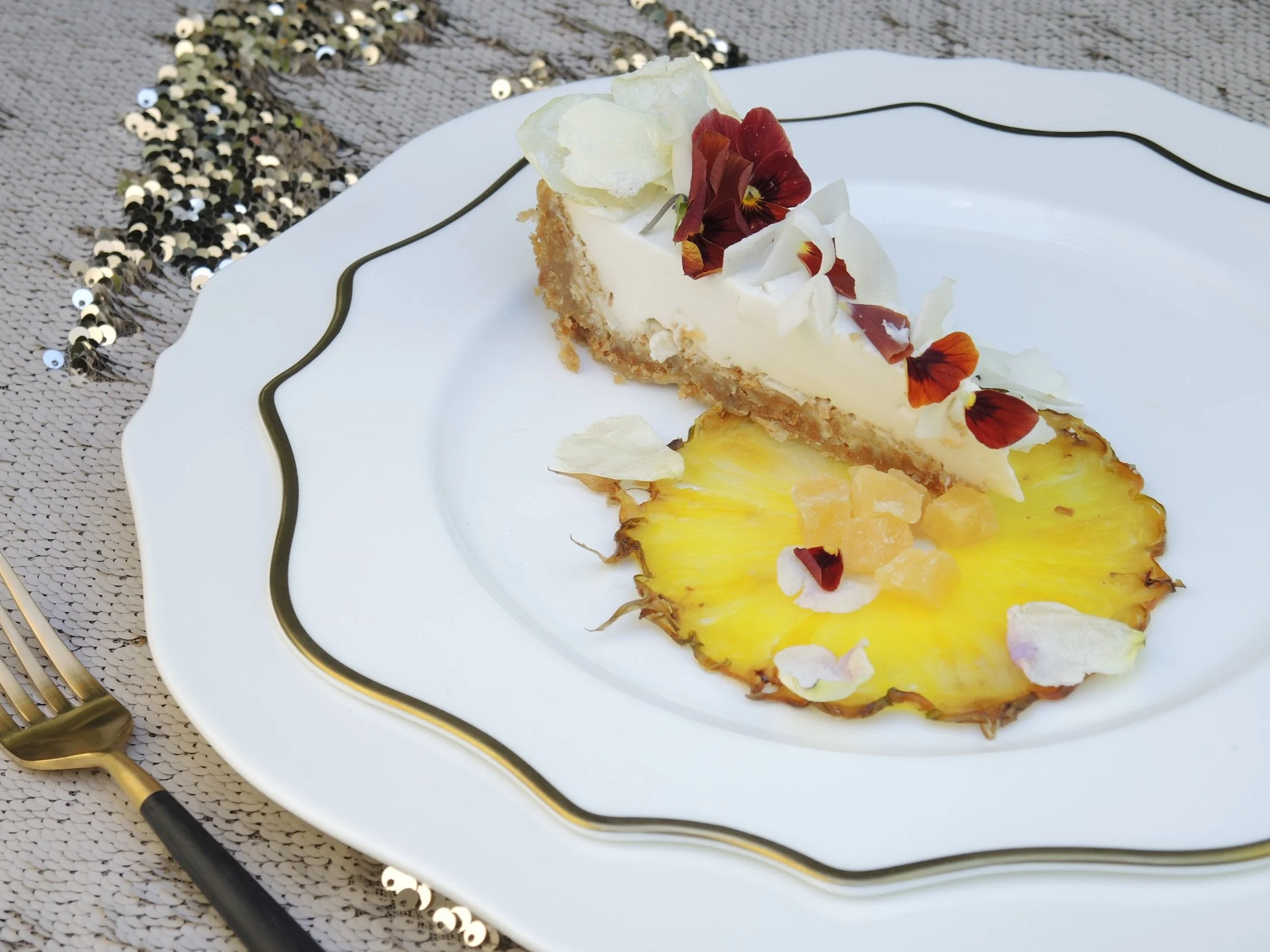 A slice of cheesecake topped with edible flowers on a white plate with gold trim, placed on a textured cream-colored tablecloth decorated with gold and black sequins, with a gold fork to the left.