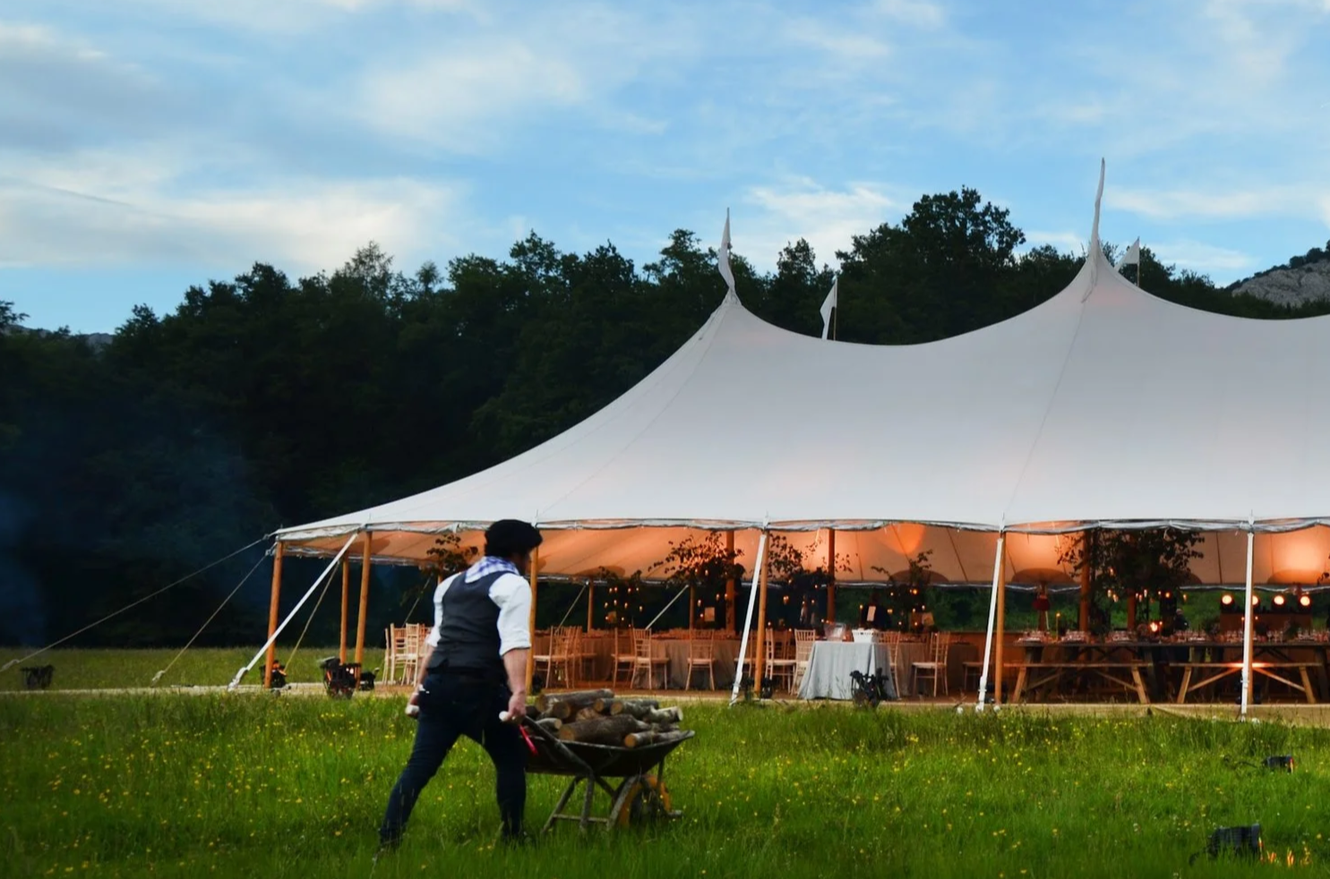 An outdoor event setup under a large white tent with string lights, surrounded by greenery and mountains in the background during evening.