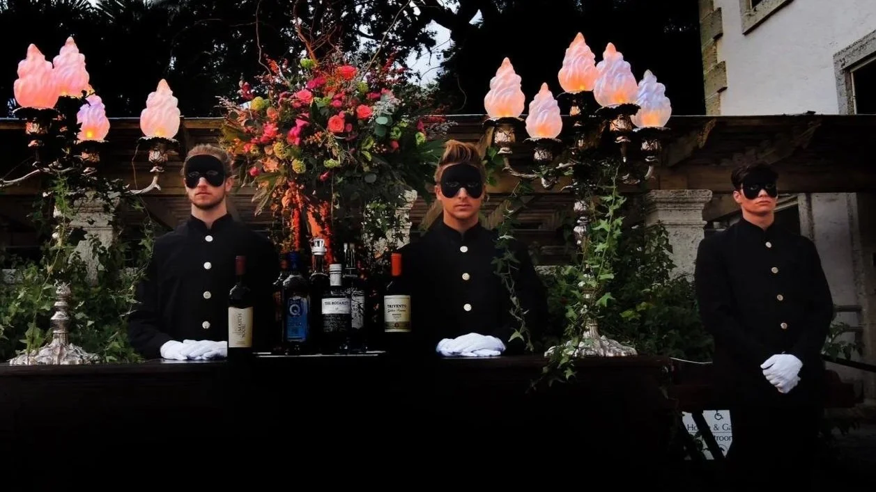 Four individuals dressed in black uniform with white gloves and eye masks stand behind a decorated table with bottles of alcohol and floral arrangements; they are outdoors at an event with decorative lighting and foliage.