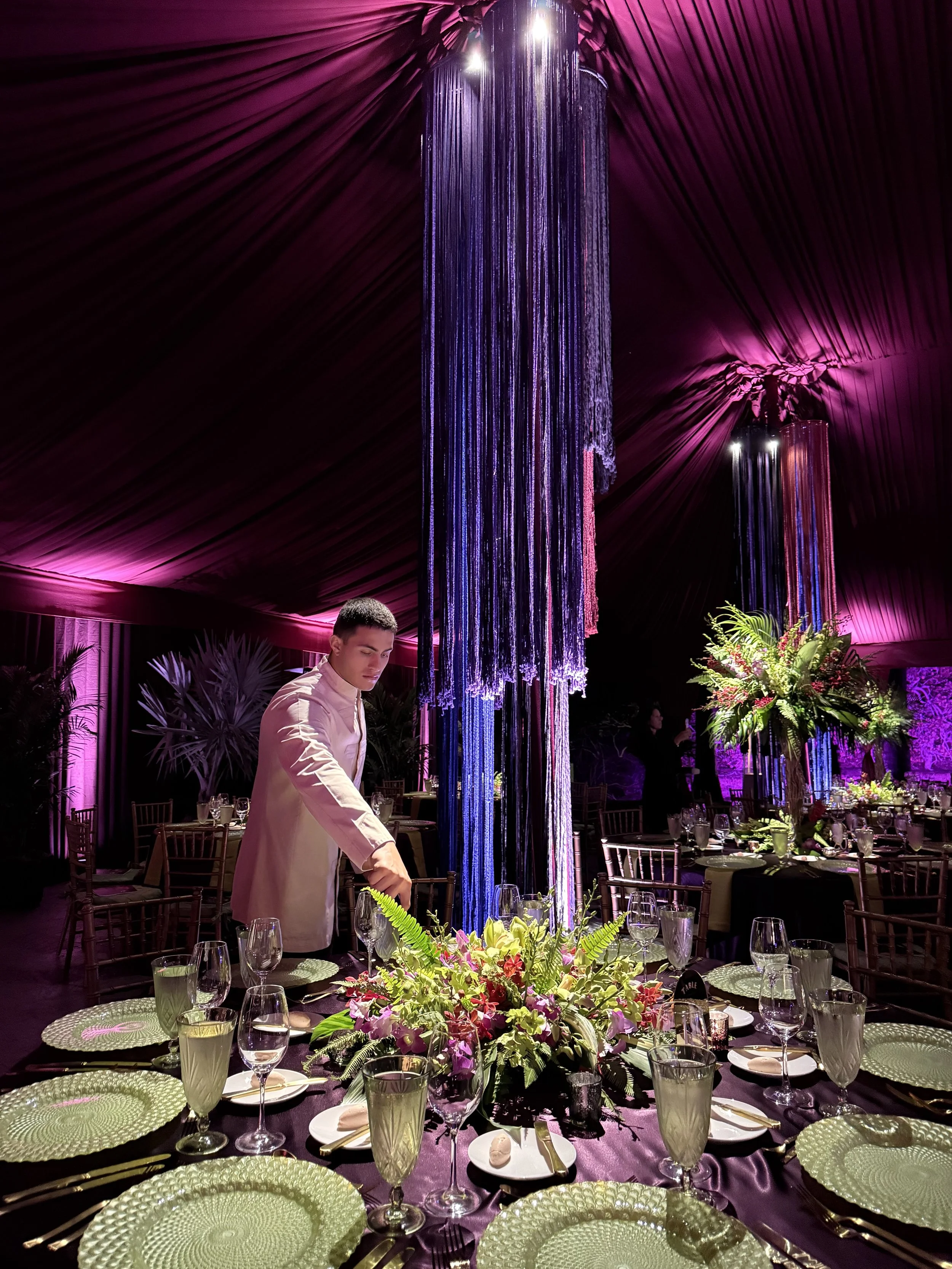 Elegant event setup with a large floral centerpiece, round tables with glassware, and a decorative hanging installation with purple and pink lighting.