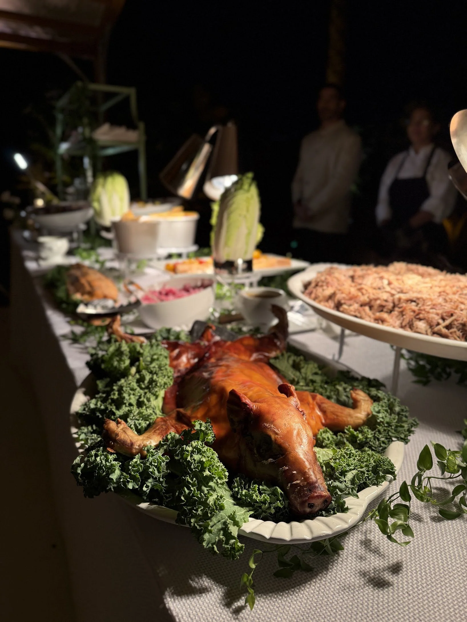 Roast pig on a large platter garnished with greens in front of a buffet table with various dishes and people in the background.