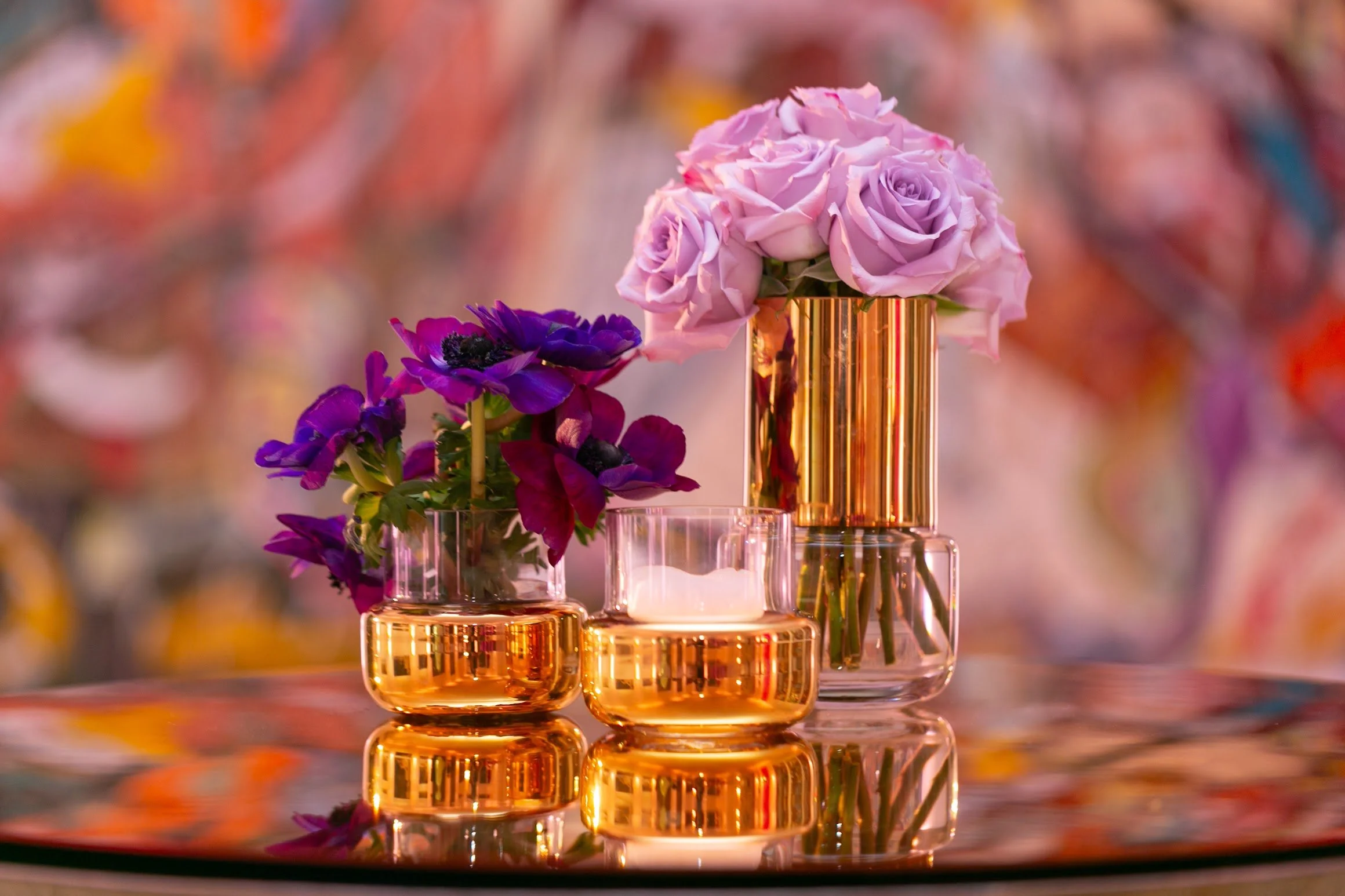 Beautiful floral arrangement with pink roses and purple anemones in gold-and-clear glass vases, reflected on a shiny surface with a colorful, blurred background.