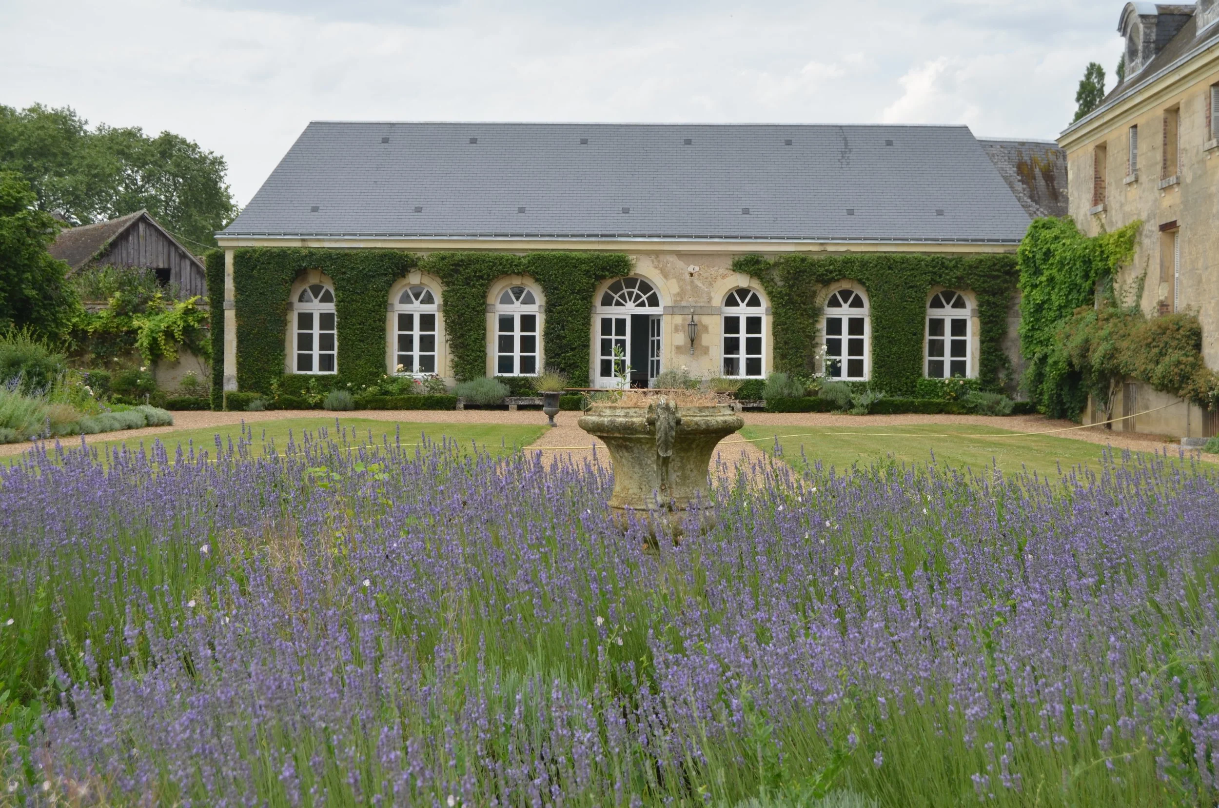 A stone building Orangerie used as a venue with large arched windows, surrounded by green ivy, with a lavender flower garden in foreground and a cloudy sky.