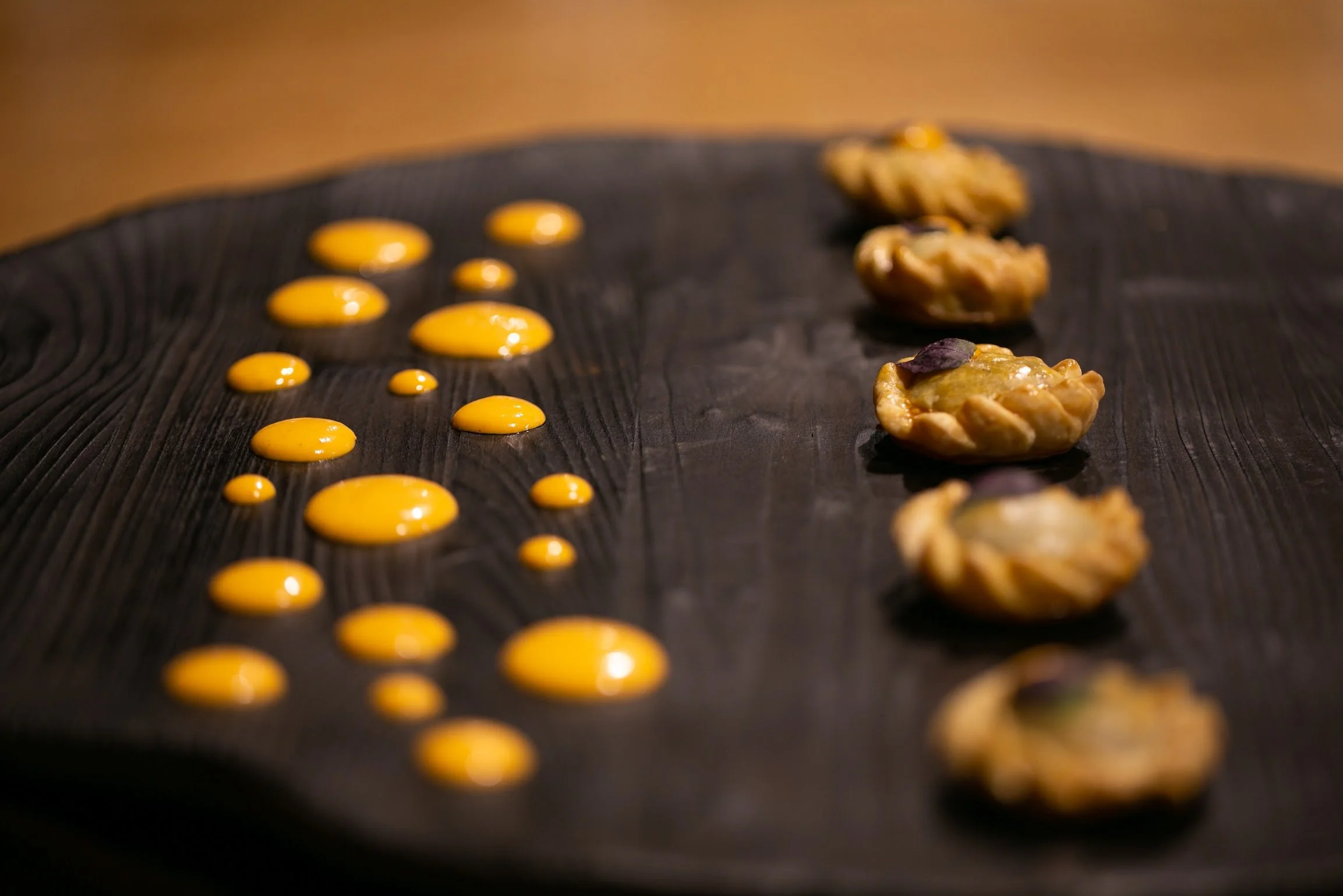 Several small pies lined up on a black surface along with dollops of yellow sauce or cream.